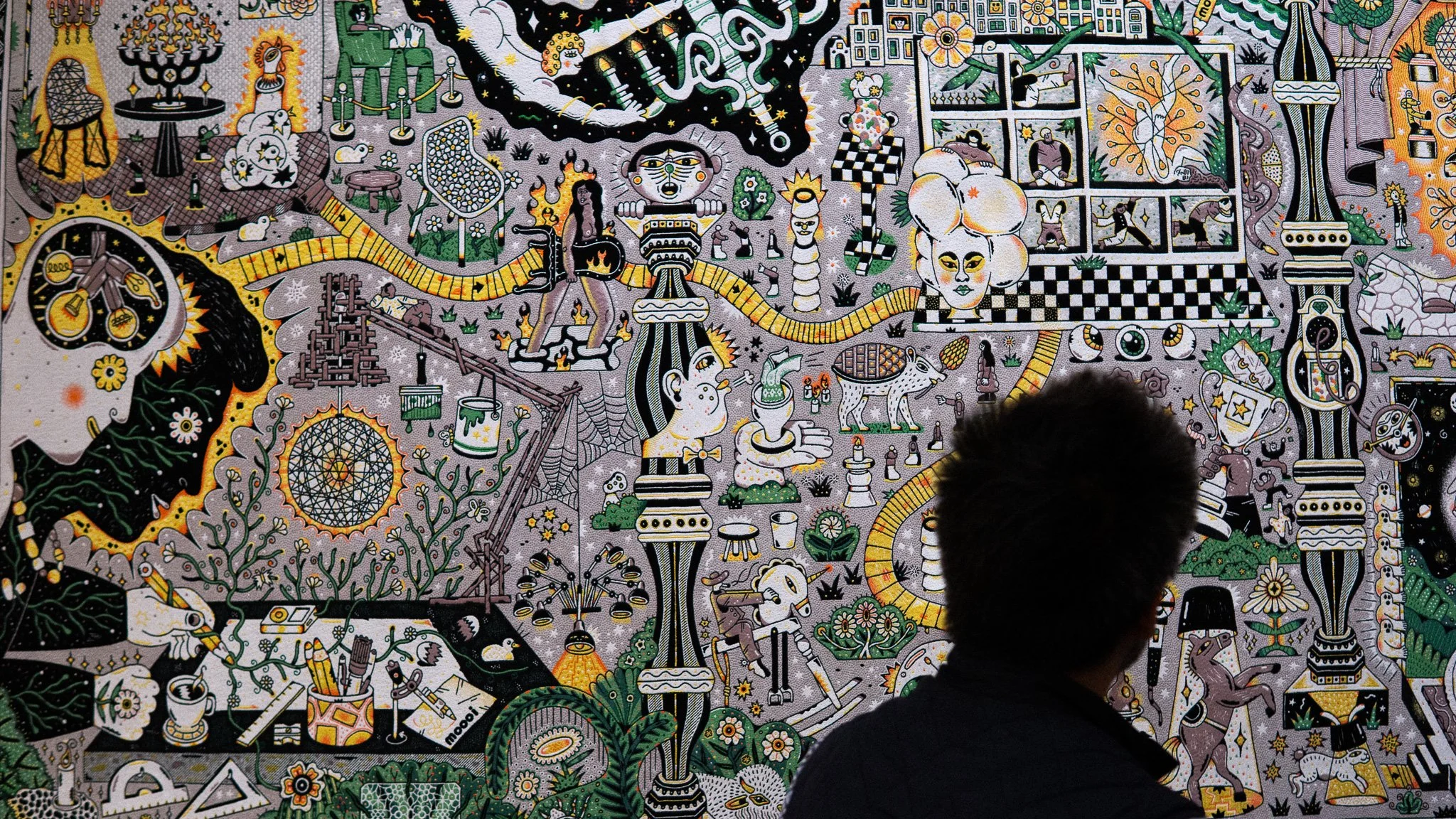 A person standing in front of a large, detailed mural with various illustrations in black, white, green, yellow, and orange, featuring abstract and whimsical designs, animals, plants, and fantastical figures.  Tapestry and jacquard weaving art