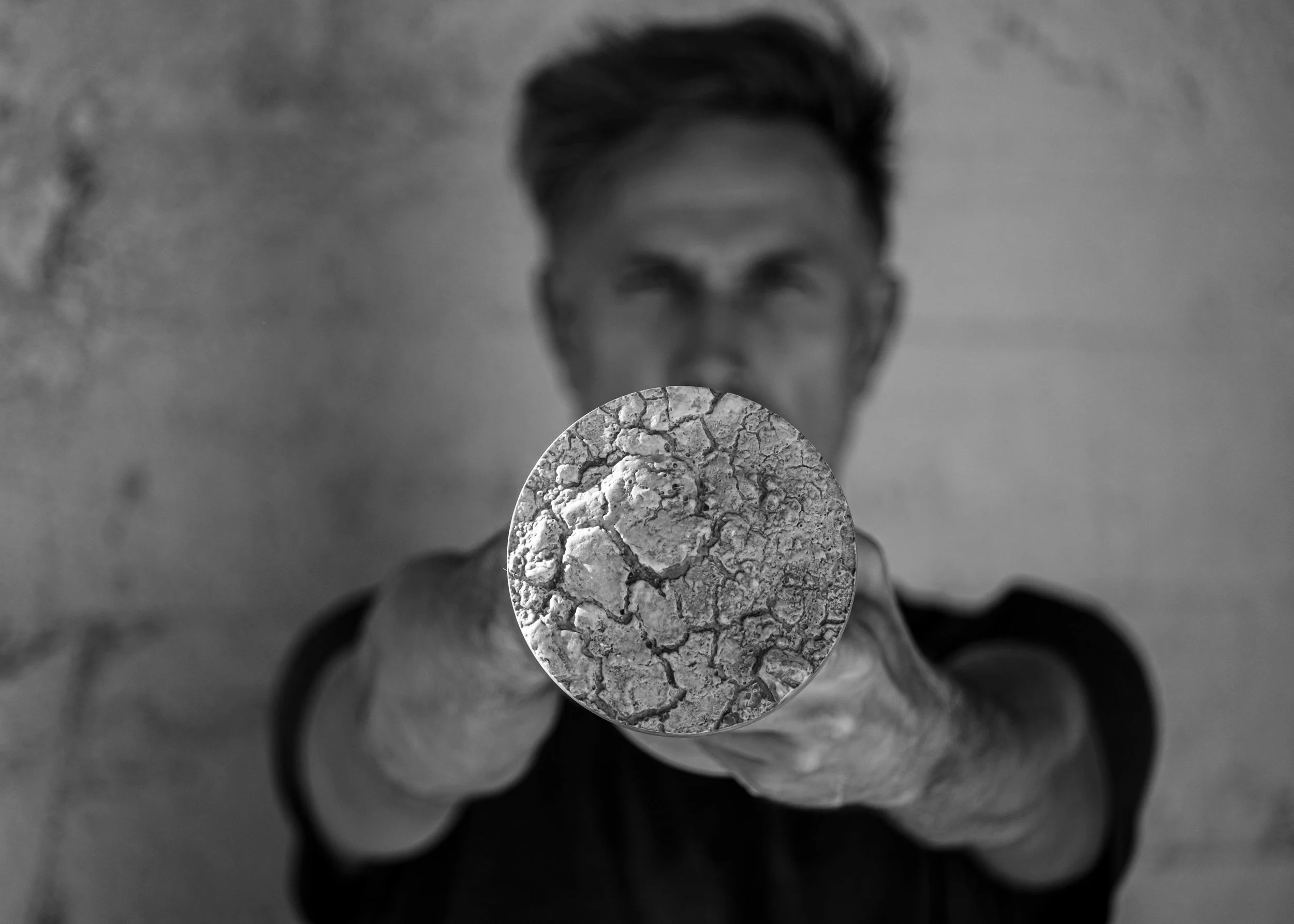A person with styled dark hair holding a round piece of cracked gray stone in front of their face, obscuring it. The background is blurred.