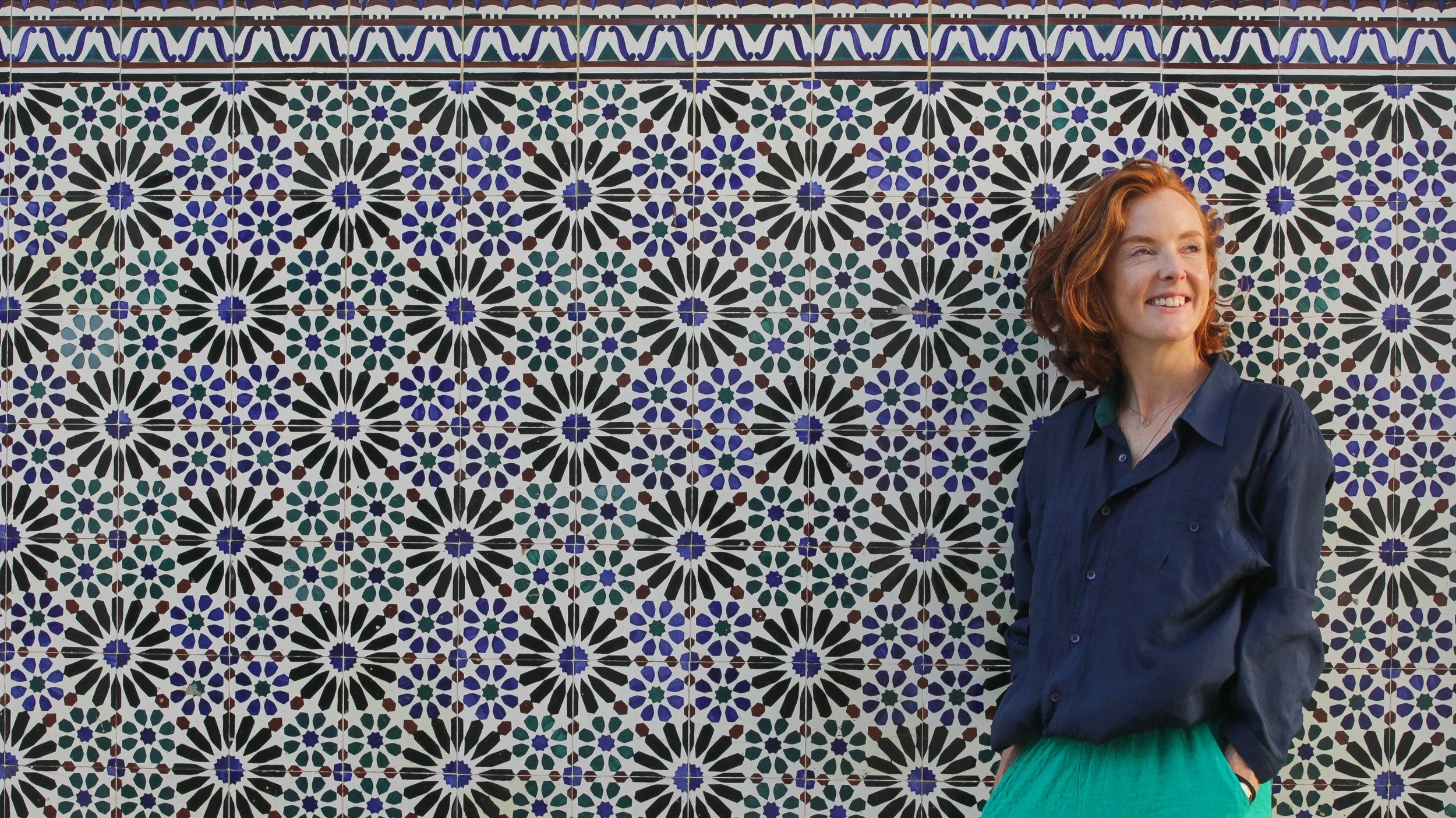 A woman with red hair, wearing a navy blue shirt and teal trousers, standing in front of a wall decorated with colorful mosaic tiles forming geometric patterns.