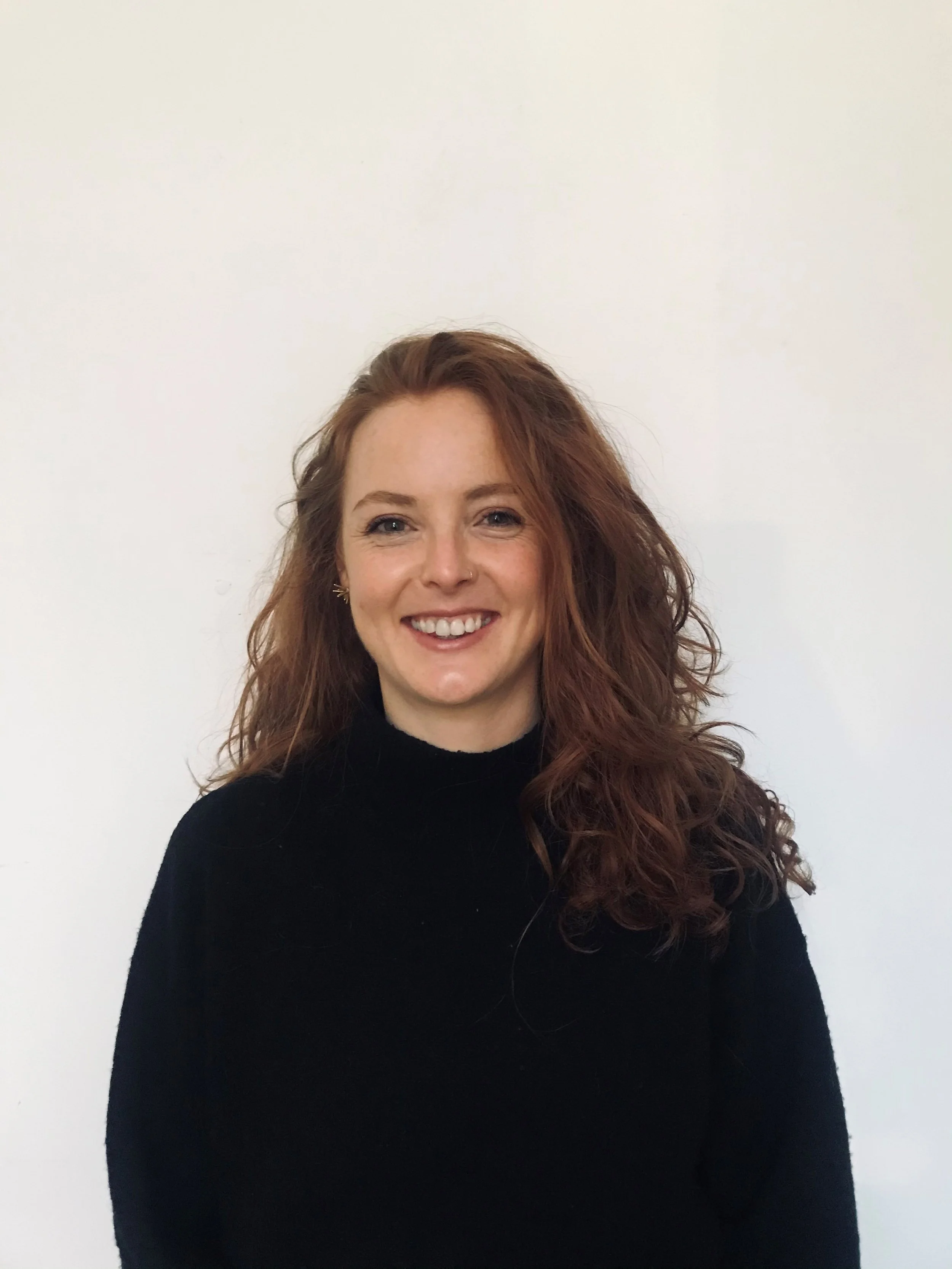 A woman with long, curly, red hair wearing a black turtleneck, smiling, standing against a plain white background.