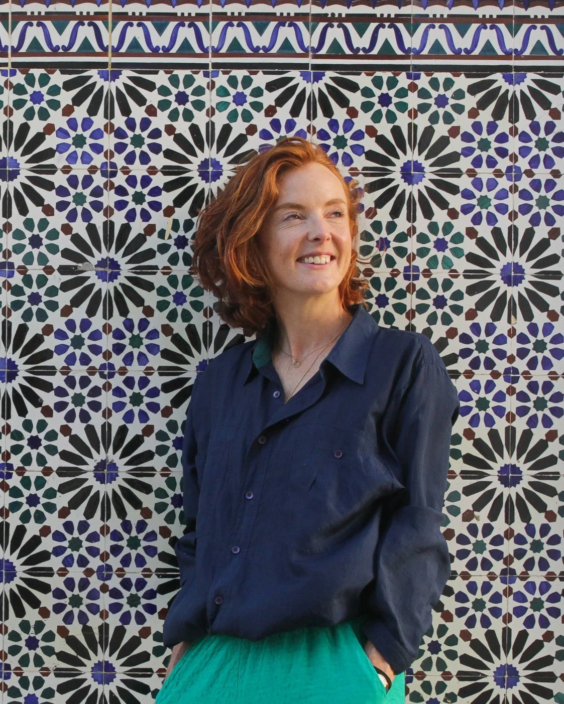 A woman with red hair smiling, standing in front of a colorful mosaic tile wall.