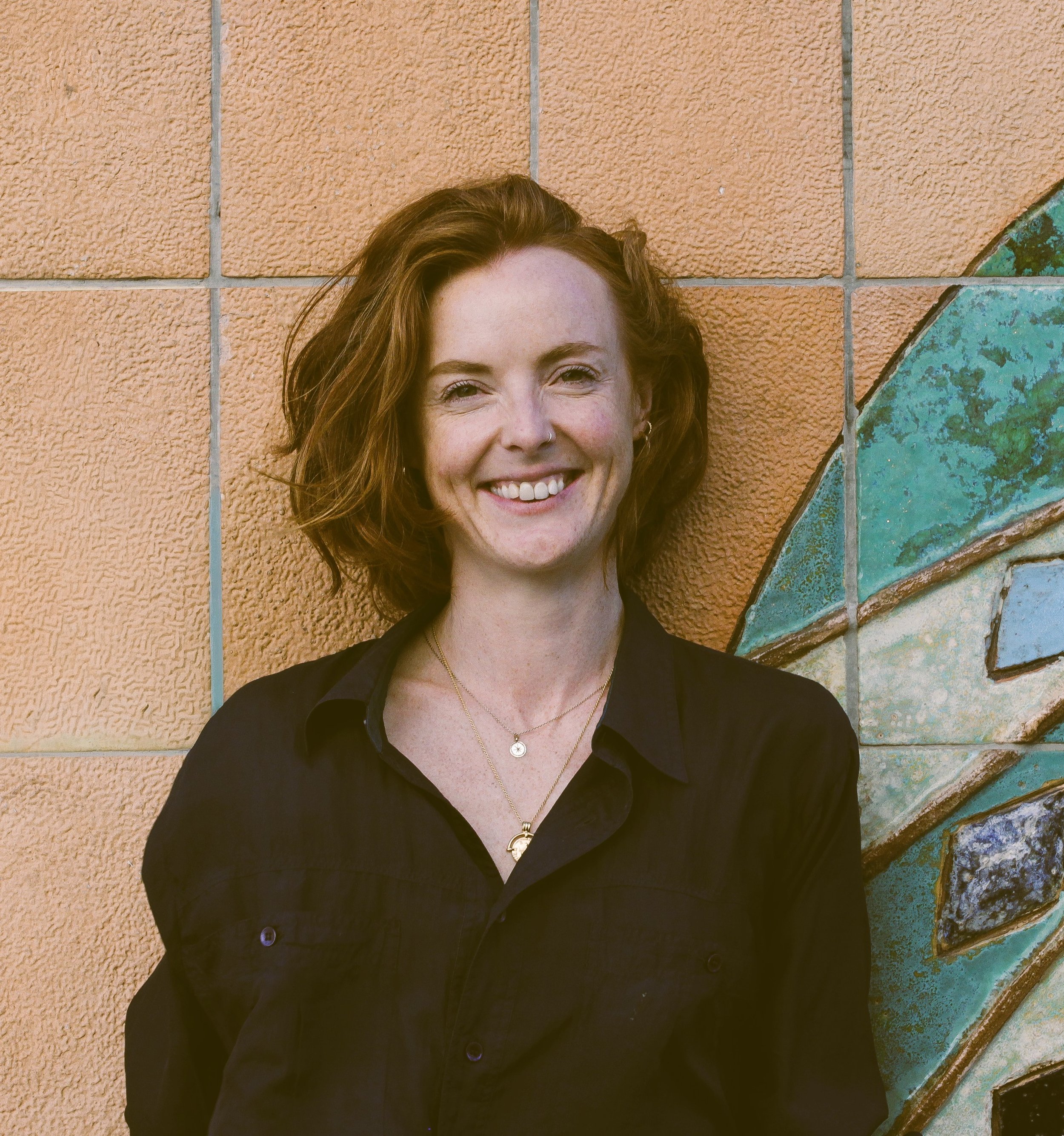 Close-up of a smiling woman with red hair, wearing a navy shirt, standing in front of an orange textured wall with ceramic artwork.