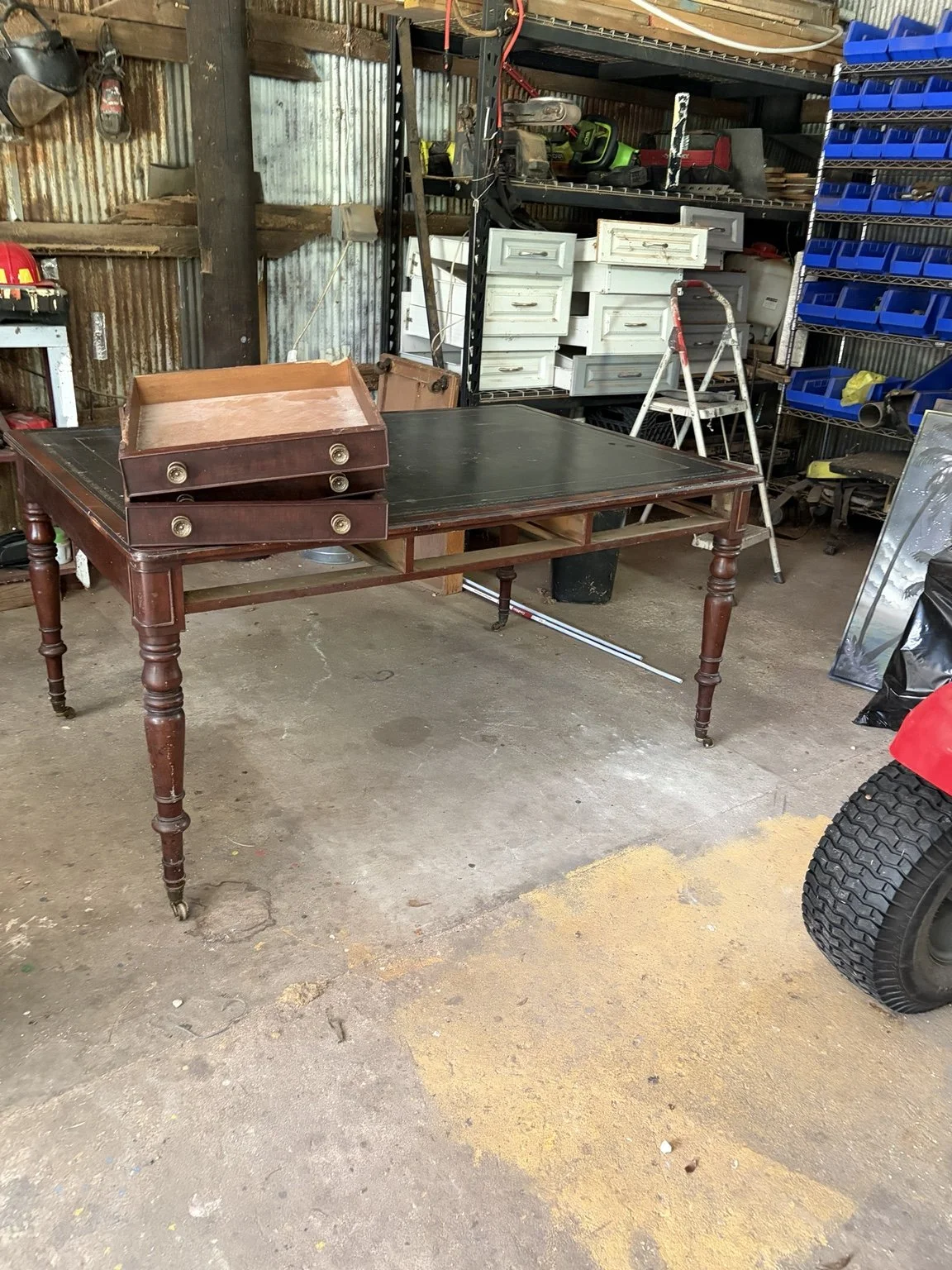 Early-1900s library table with turned wooden legs, brass feet and inlay, with its drawers removed and resting on top at the workshop.