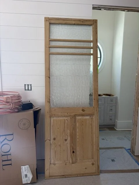 A wooden door with a glass window in the upper part and wooden panels in the lower part, leaning against a wall in a room under construction.