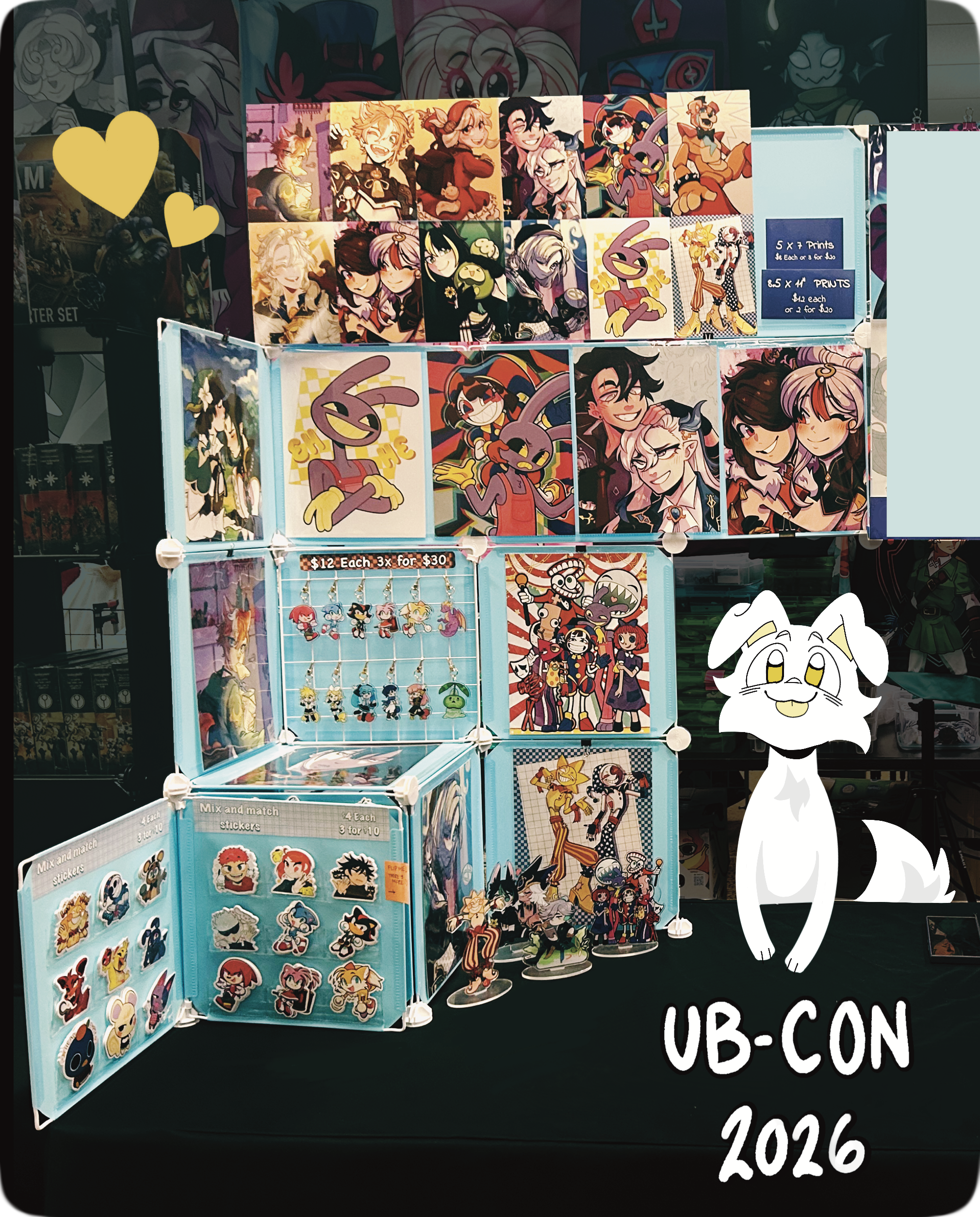 Display of colorful stickers, prints, and keychains of various cartoon and video game characters arranged on a multi-tiered stand at a booth, with a handwritten sign indicating prices. A cartoon white dog illustration is visible in the bottom right corner.