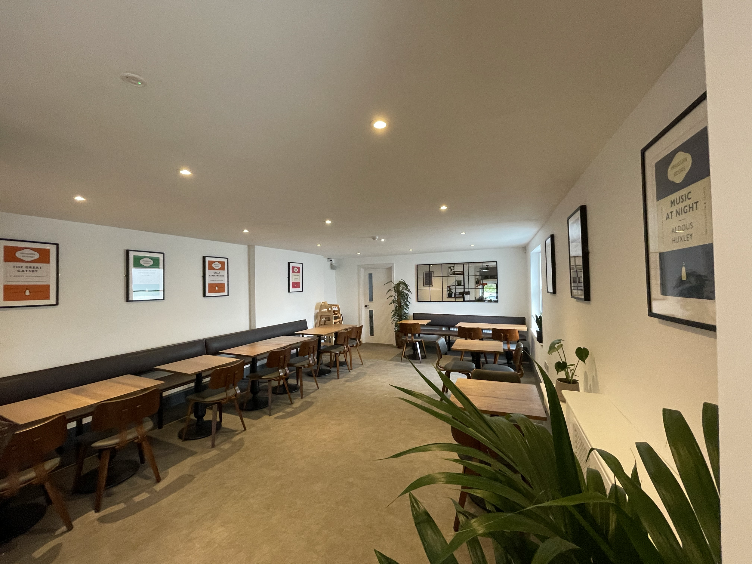 A cozy cafe or lounge area with wooden tables and chairs along a white wall decorated with framed posters. There are additional seating options along the wall, a large window letting in natural light, and some indoor plants adding greenery to the spa