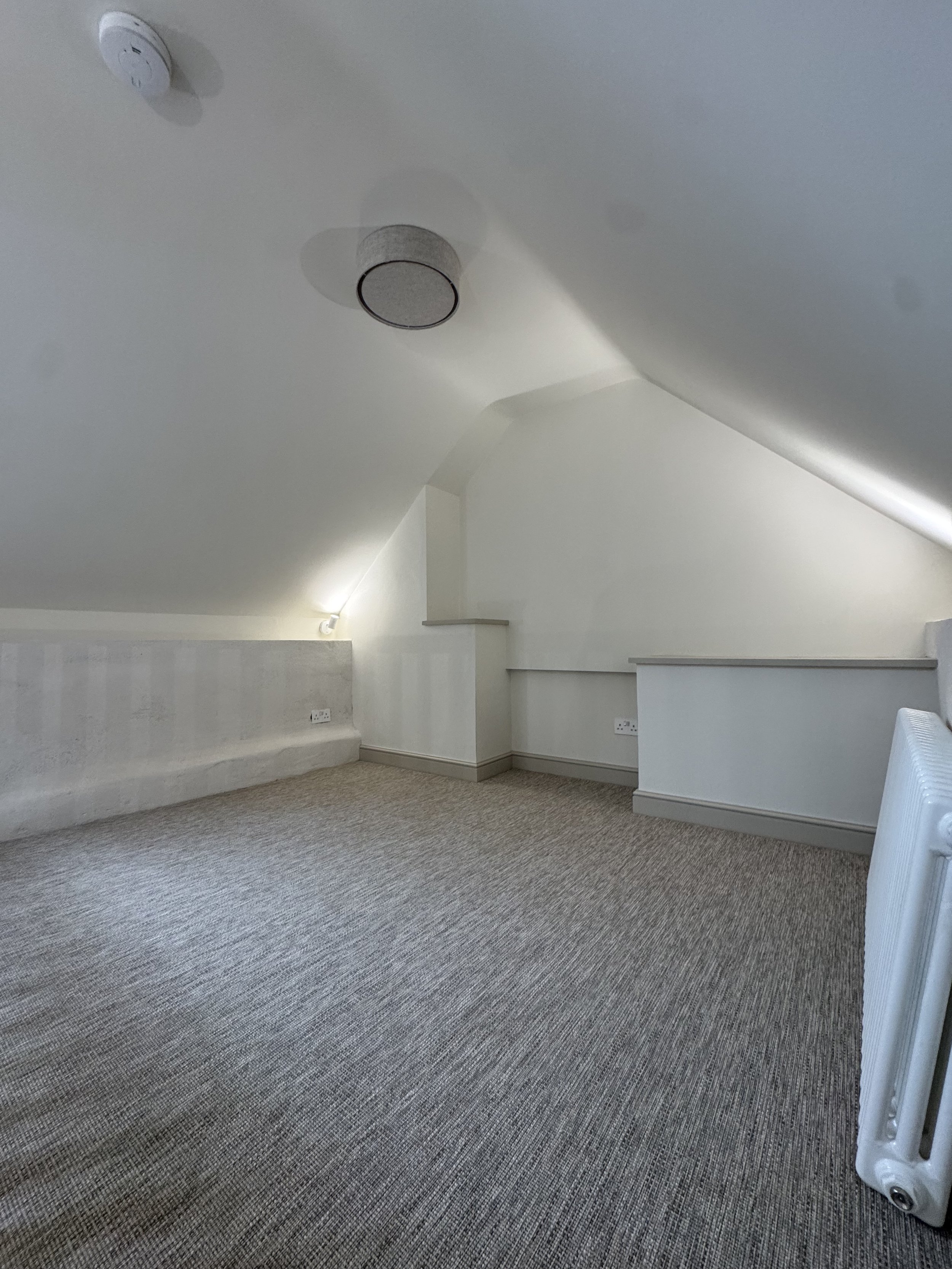 A corner of a room with a carpeted floor, a white radiator, and a sloped ceiling. There are electrical outlets on the wall, and a smoke detector and a ceiling speaker are visible on the ceiling.