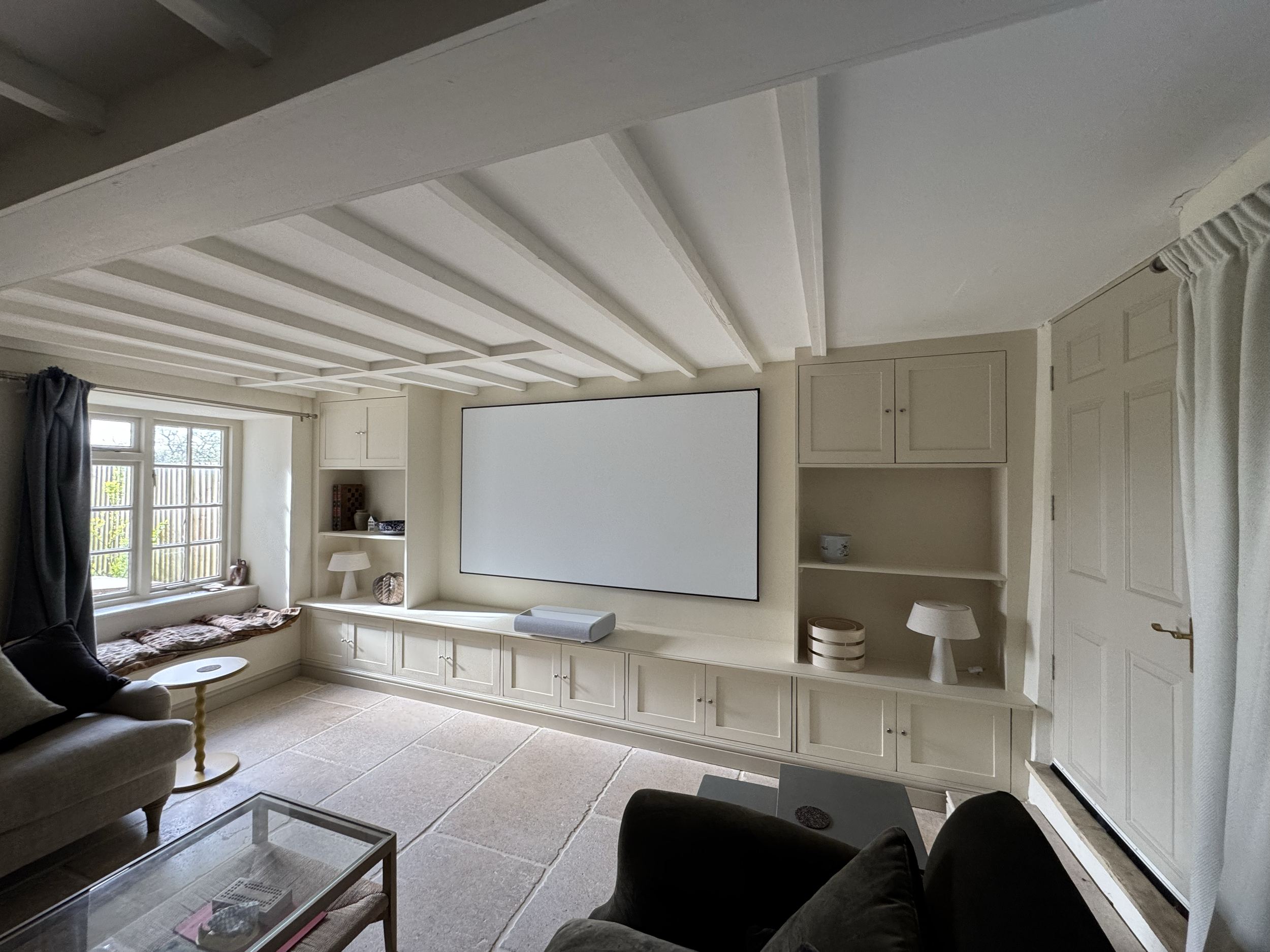 Living room with beige walls and a white ceiling with beams, a large window with curtains, a gray sofa, a beige armchair, a small lady table, built-in white cabinets, a large wall-mounted white screen, and a tiled floor.