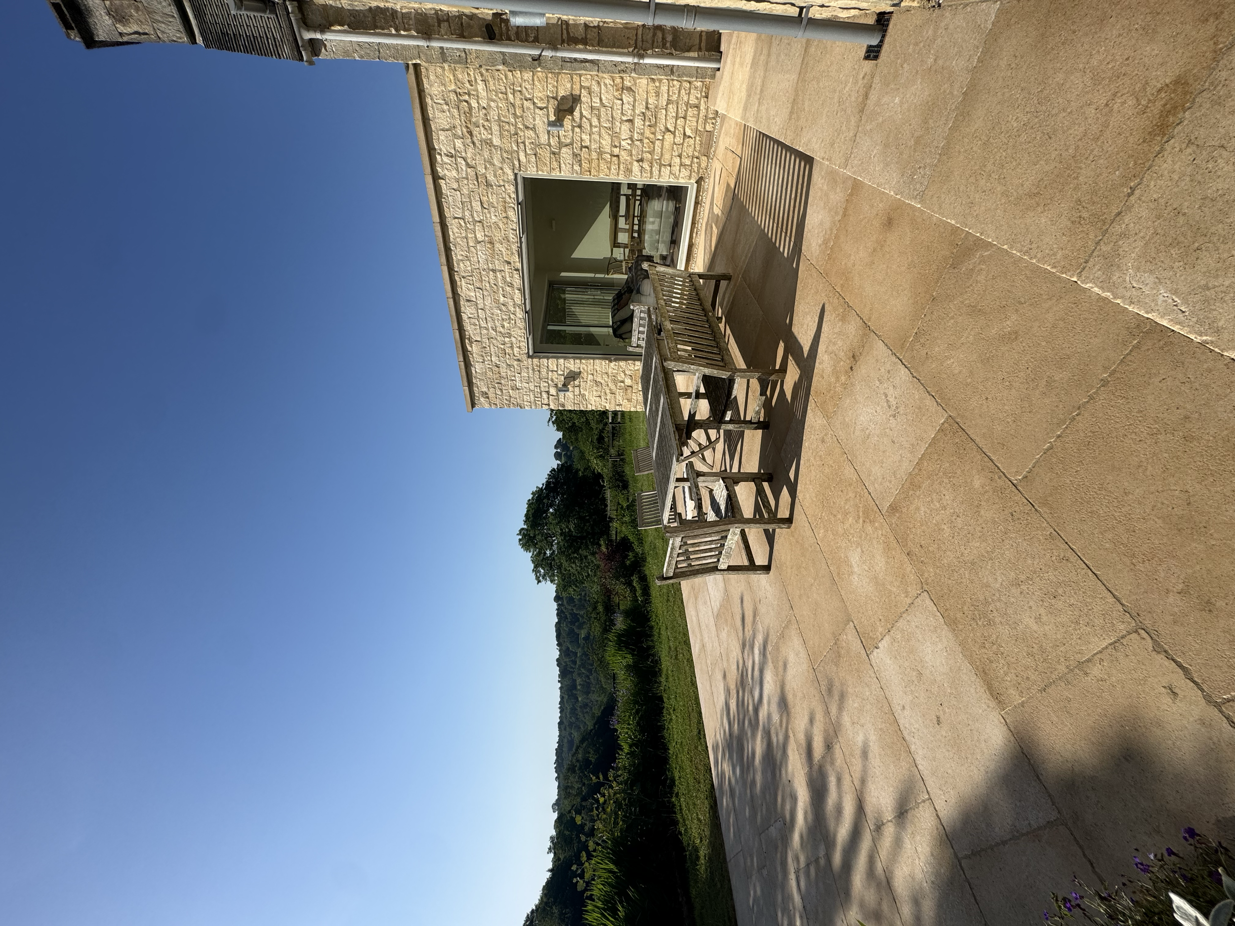 The image shows an outdoor patio area with a wooden table and chairs, stone flooring, a stone building wall with a large window, and green trees in the background under a clear blue sky.