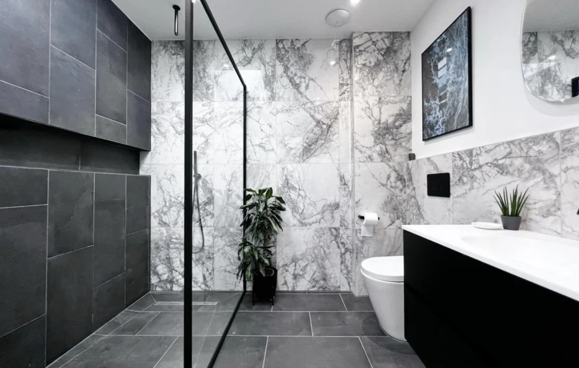 Modern bathroom with gray and white marble walls, black and gray tiled floor, a glass shower enclosure, a white toilet, a black vanity with a white countertop, a small plant, a wall mirror, and a framed artwork.