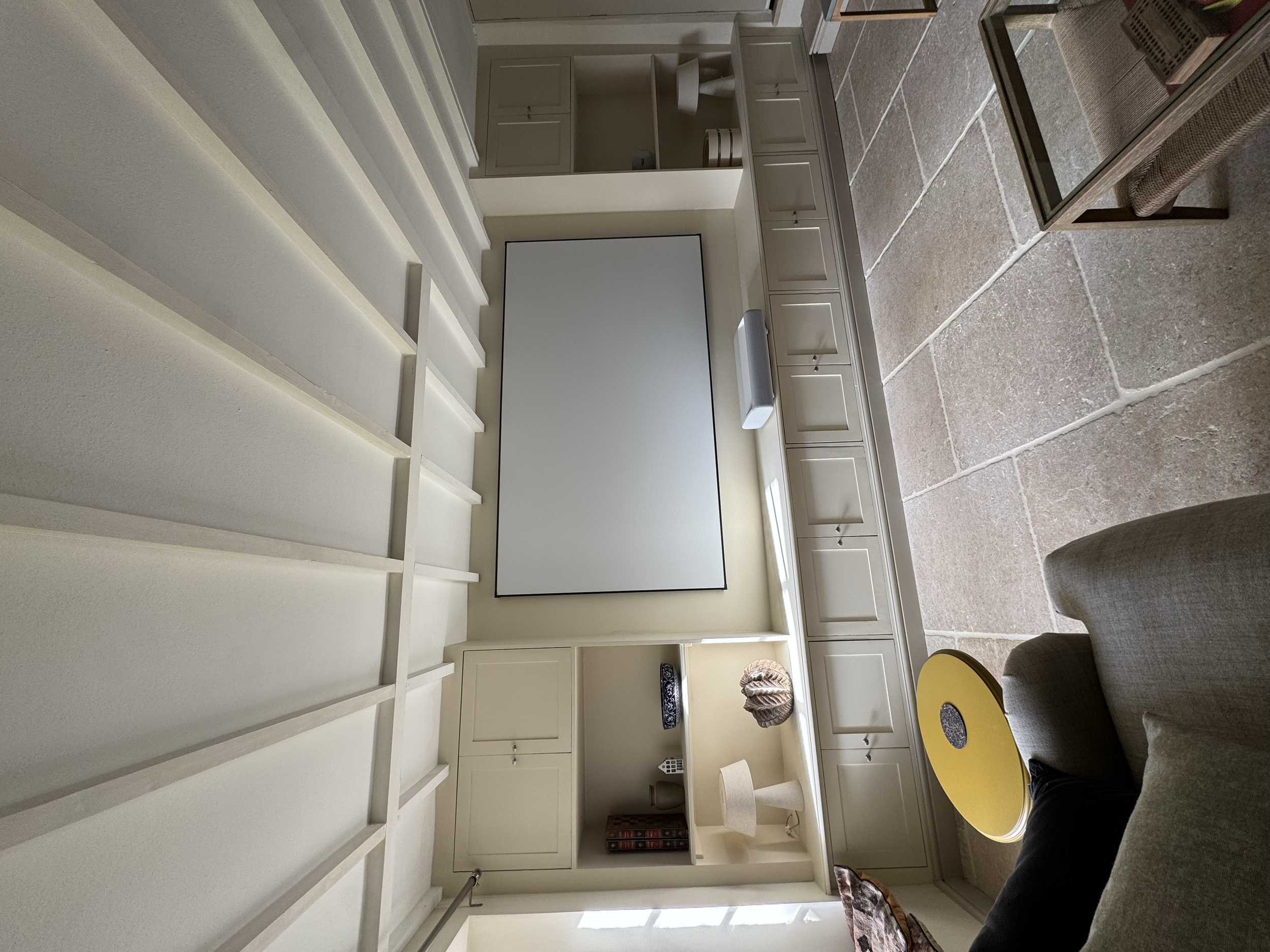 Living room with built-in white shelves, a tv, and beige cabinetry. There is a stone-tiled floor, a yellow coffee table, and a gray sofa. Part of a person is visible sitting on the sofa.