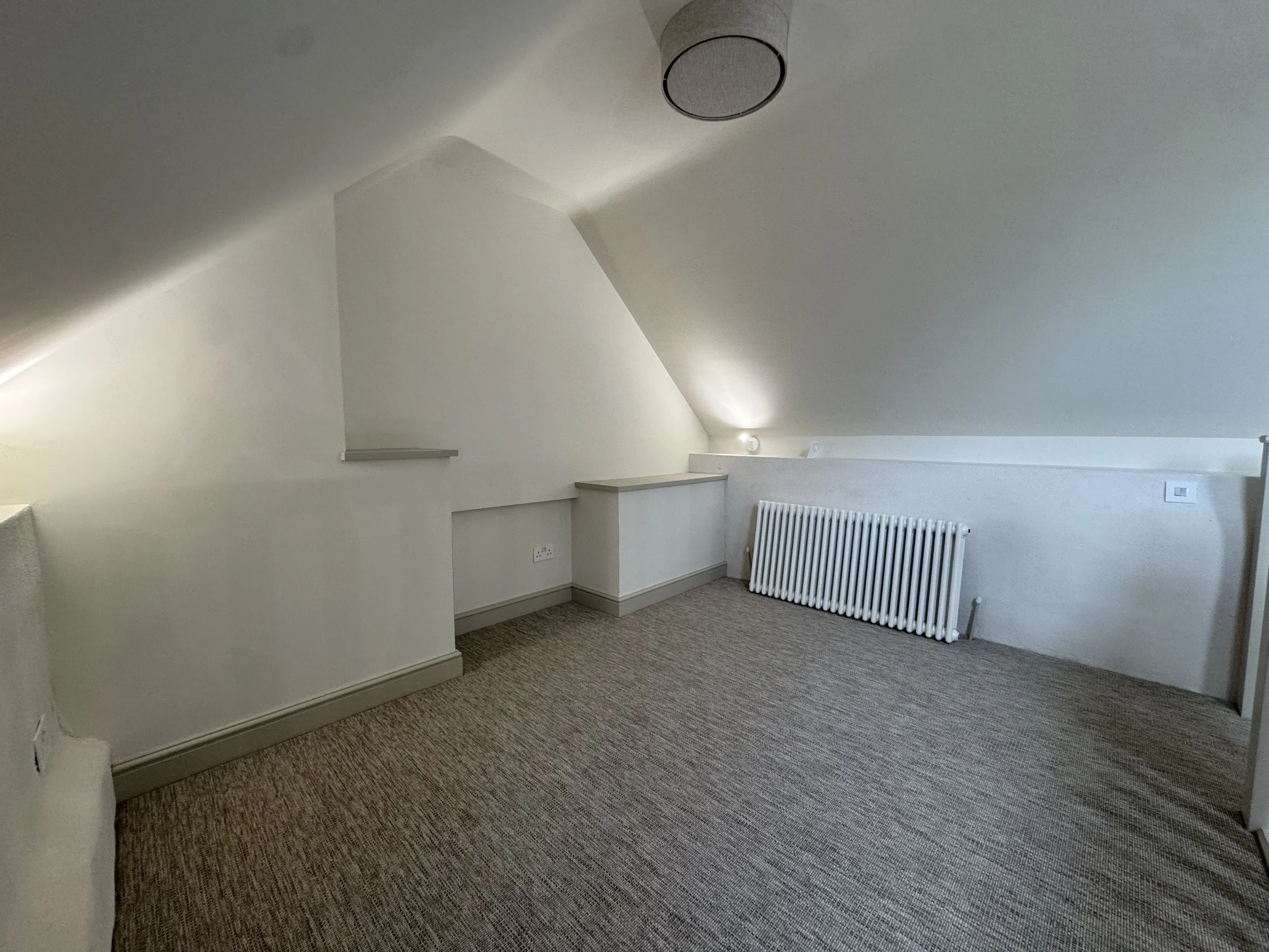 A minimalist attic room with a sloped ceiling, white walls, beige carpet flooring, a white radiator, and a small built-in shelf. A ceiling light fixture is visible, and there are multiple electrical outlets on the walls.