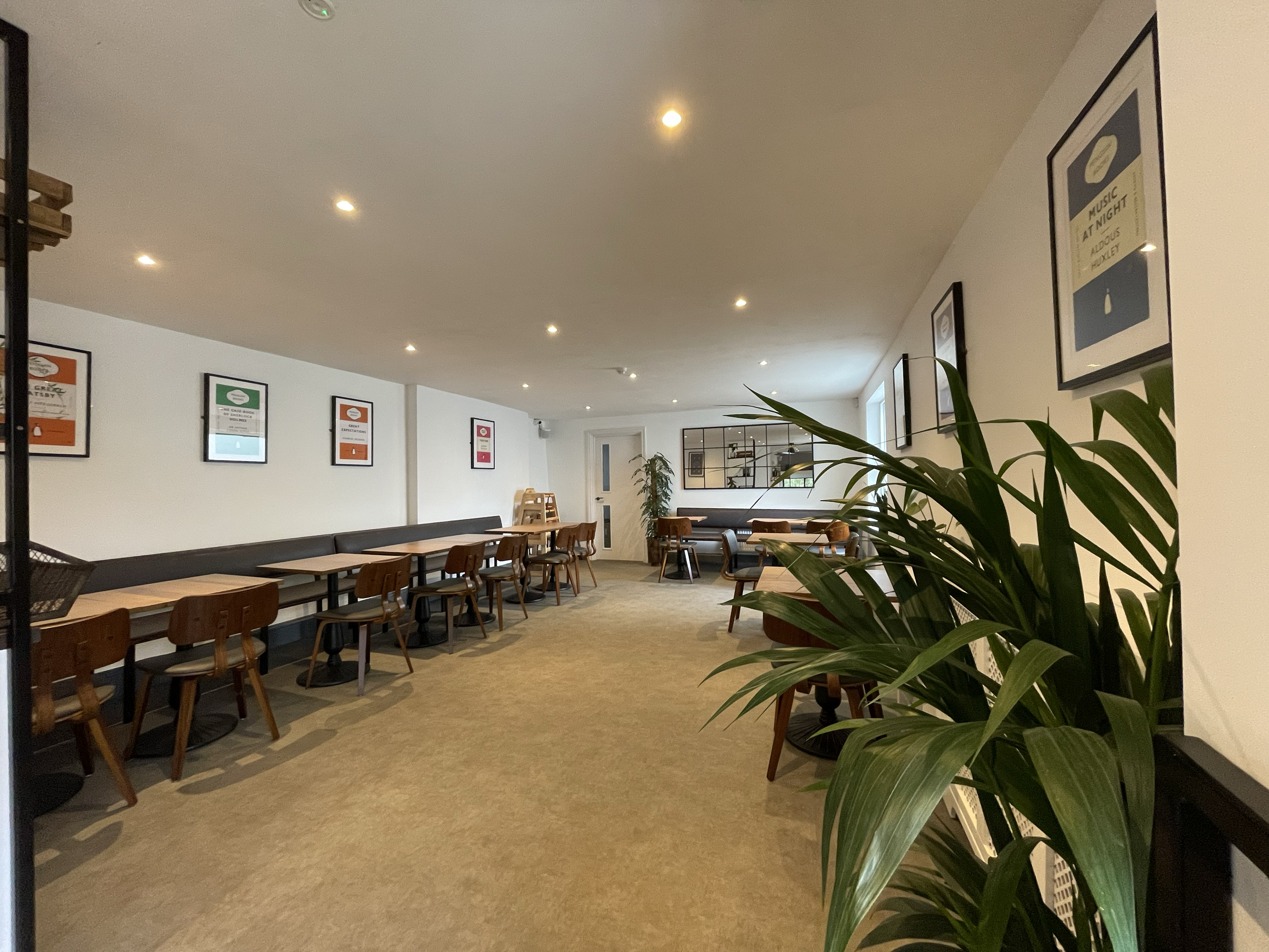 An interior view of a cafe or restaurant with wooden tables and chairs, framed posters on the white walls, a large mirror, and green plants near the window, illuminated by ceiling lights.