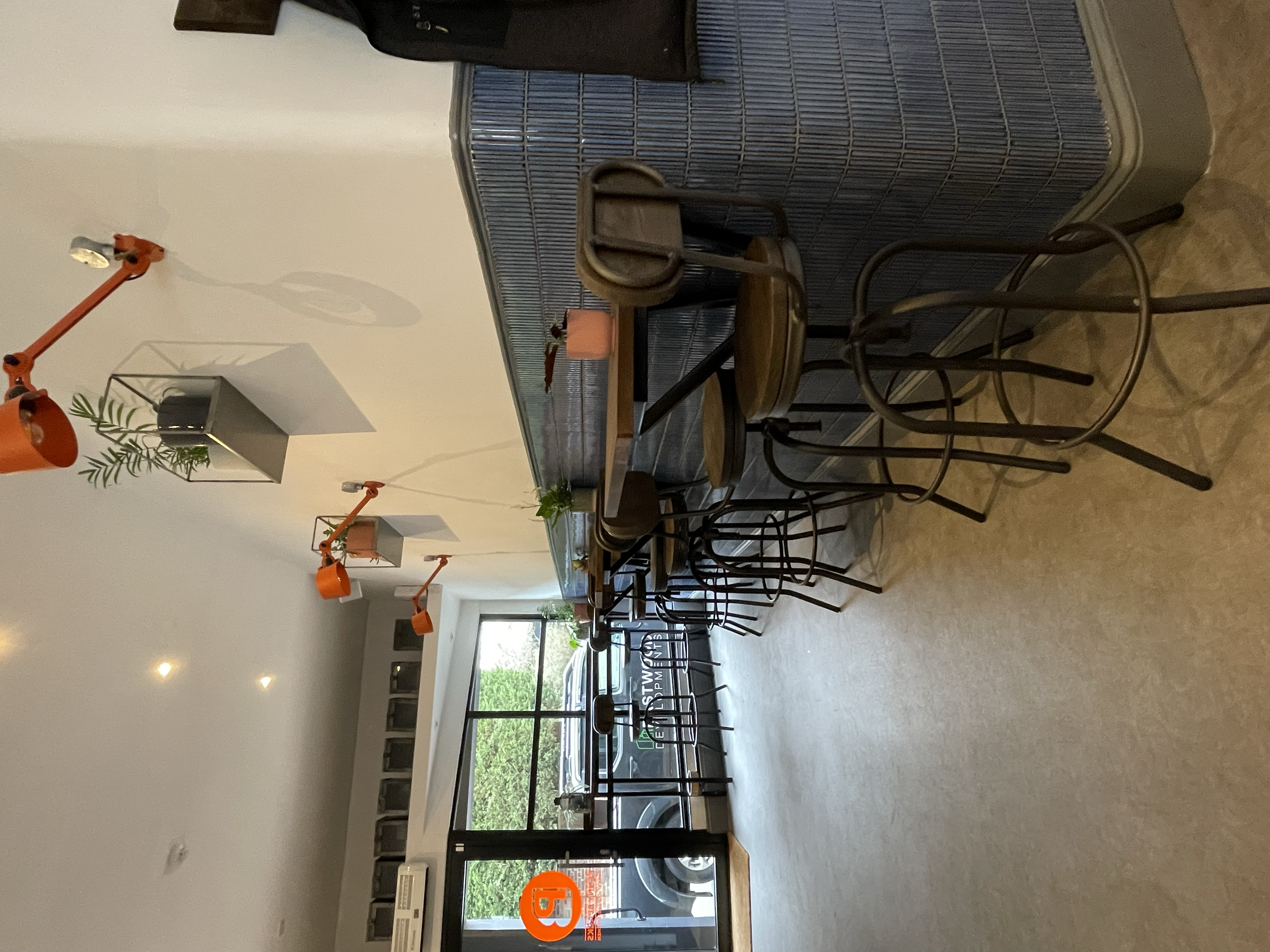 Interior of a modern cafe or restaurant with black chairs and tables, large windows, and orange lighting fixtures on the ceiling.