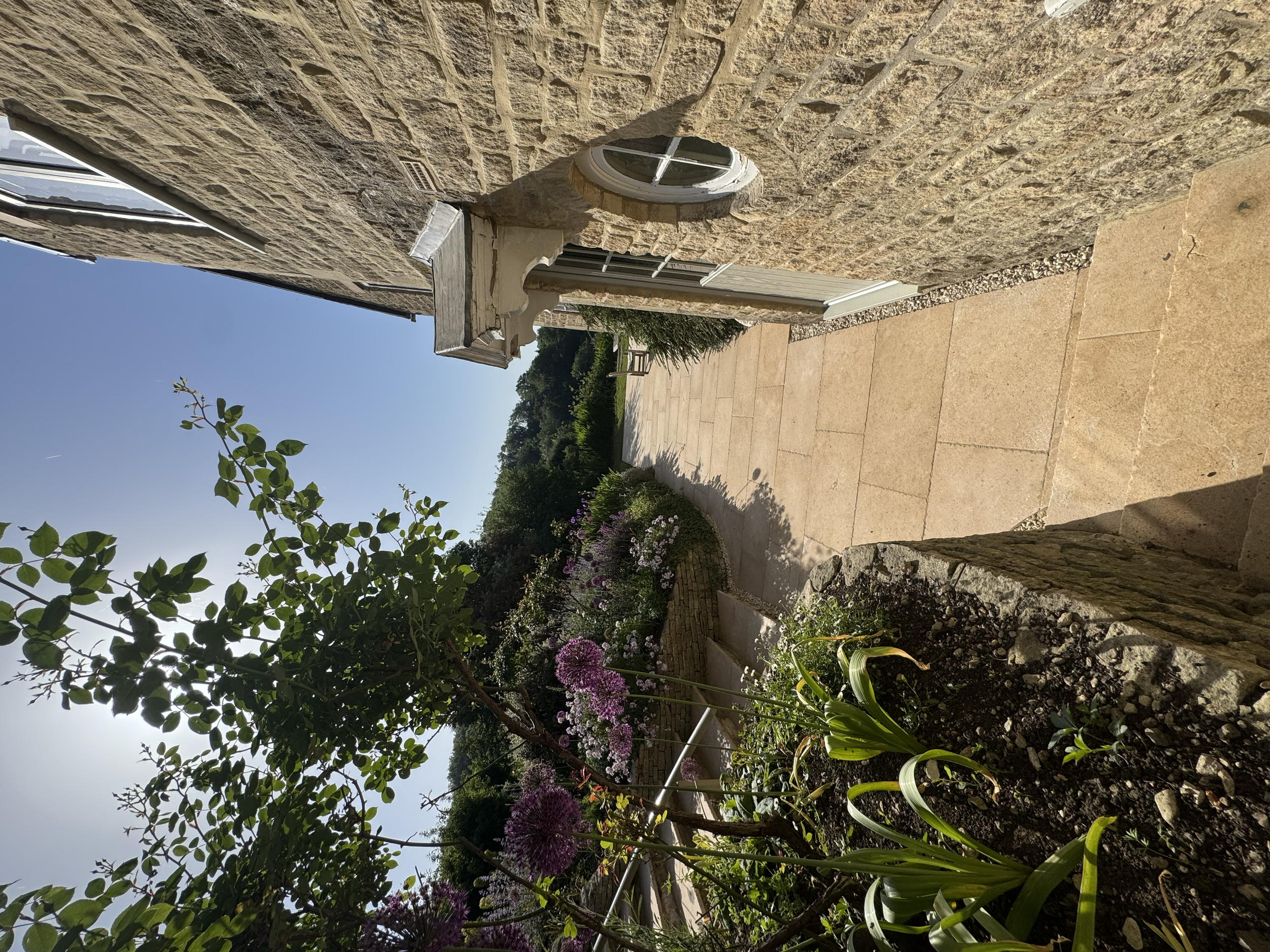 A stone wall with a circular window, a concrete planter, a white doorway, a garden with purple and pink flowers, green trees, and a paved pathway.