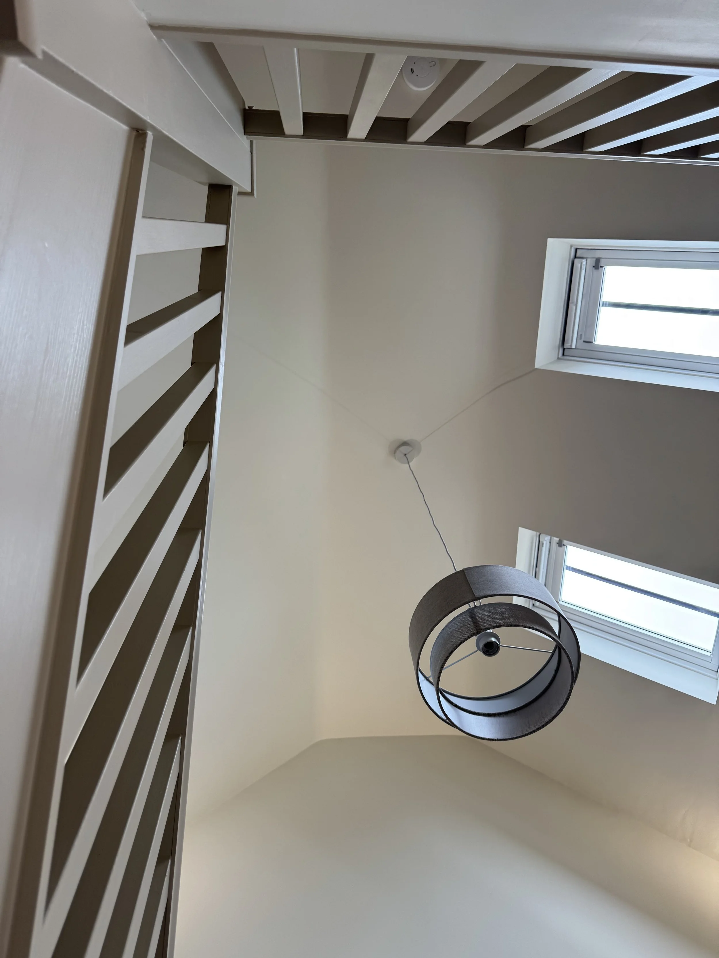 Interior view of a room with a ceiling light fixture, two windows, and a stair railing, taken from the bottom looking up.