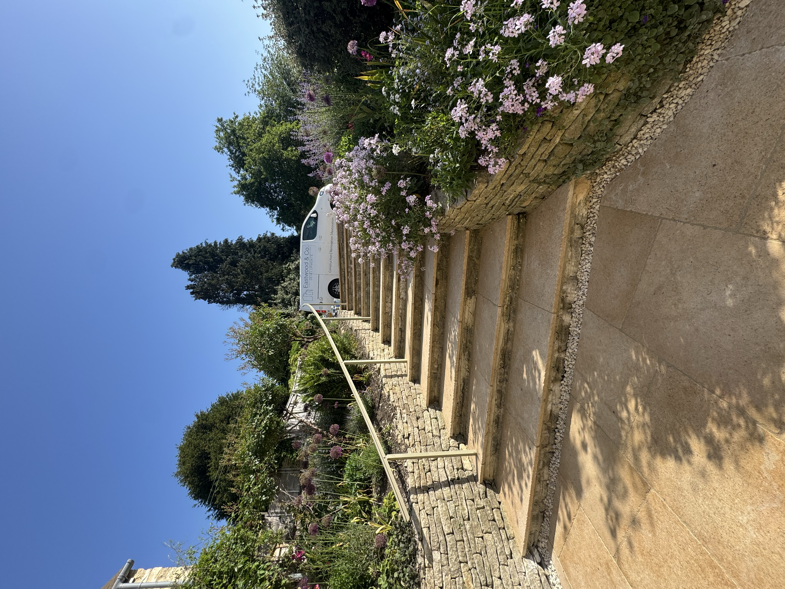 A garden with stone steps leading up to a higher level, bordered by a flower bed with pink and purple flowers. A white van is parked in the background near a sign that reads "Eastwood & Co. Building Renovations". Green trees and a clear blue sky are 