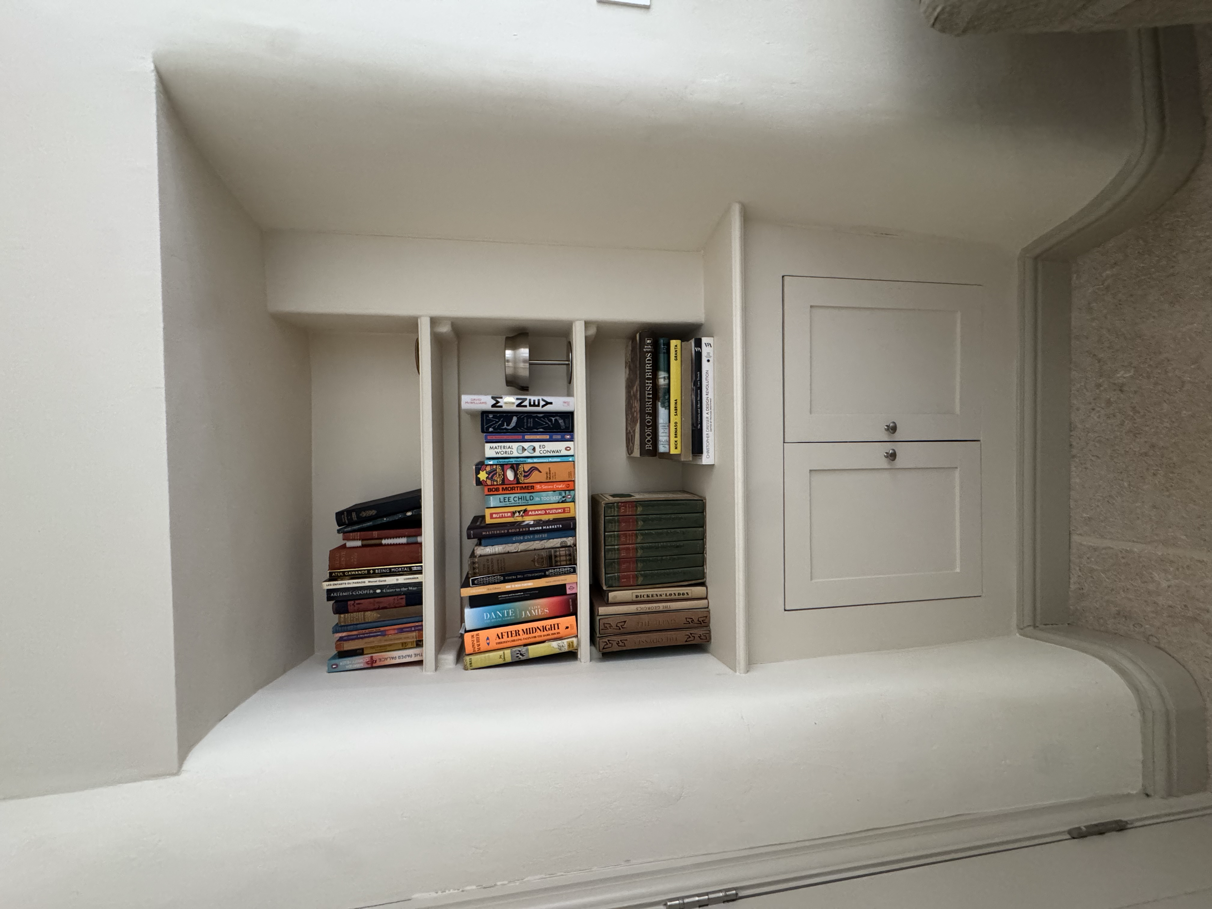 White built-in bookshelf with two shelves filled with books, located within a white wall nook next to a door with silver knobs, covered with beige carpet.