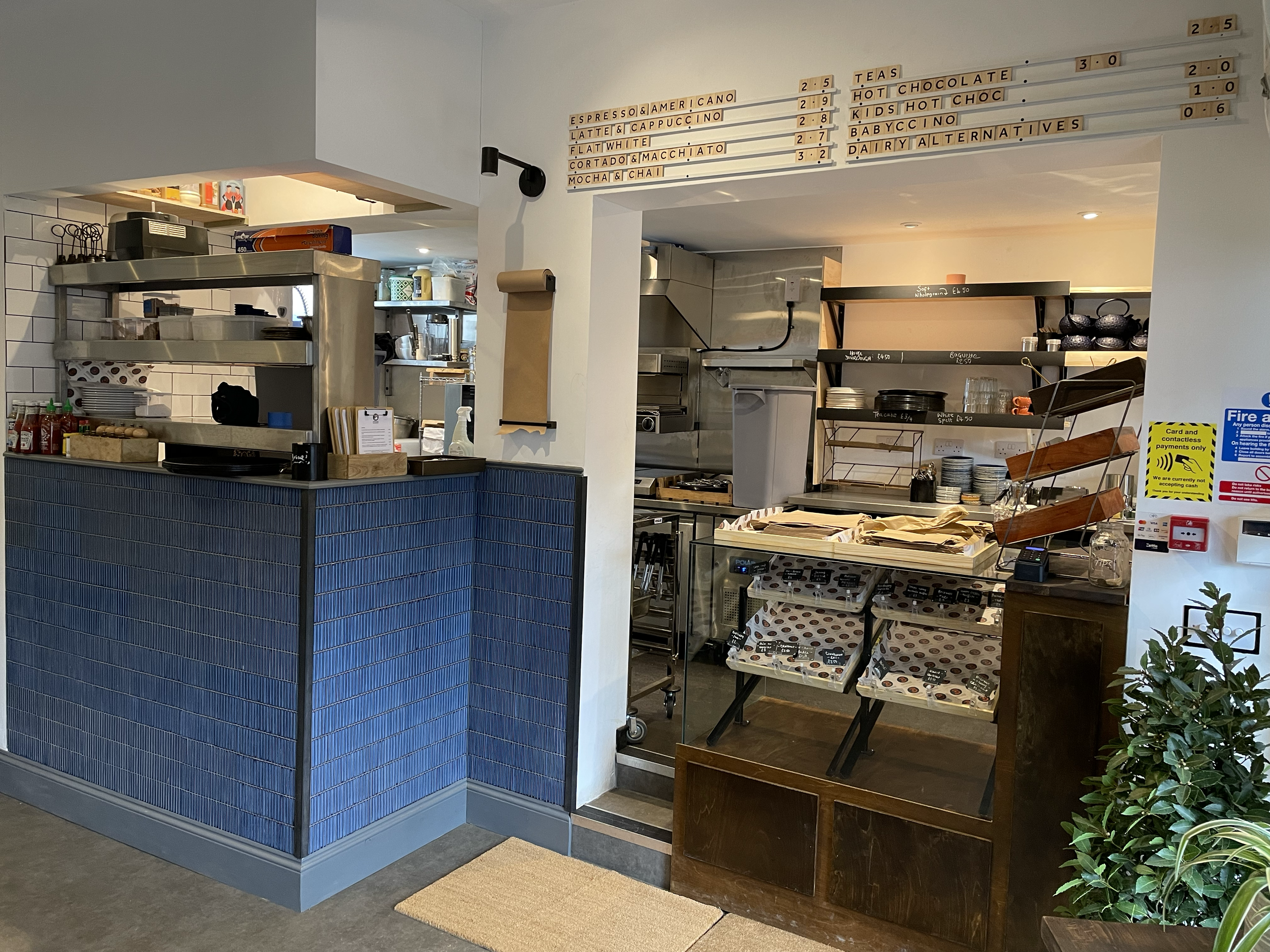 Interior view of a cafe or restaurant counter area with menu board and shelves of supplies and dishes, featuring a blue tiled front counter and warm lighting.