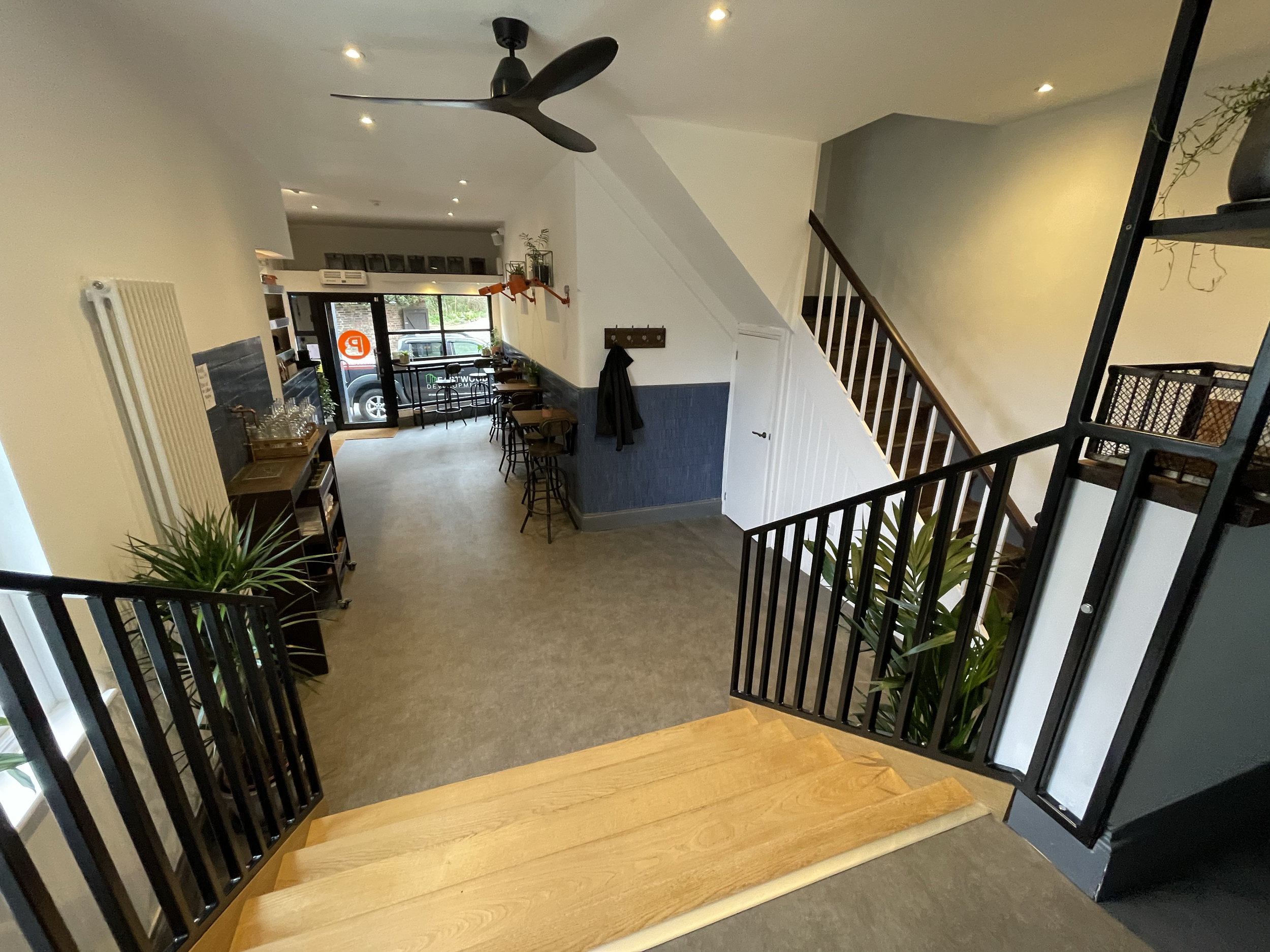 View of a staircase with wooden steps and black metal railings, overlooking a cozy commercial space with seating, plants, and a large window at the front entrance.