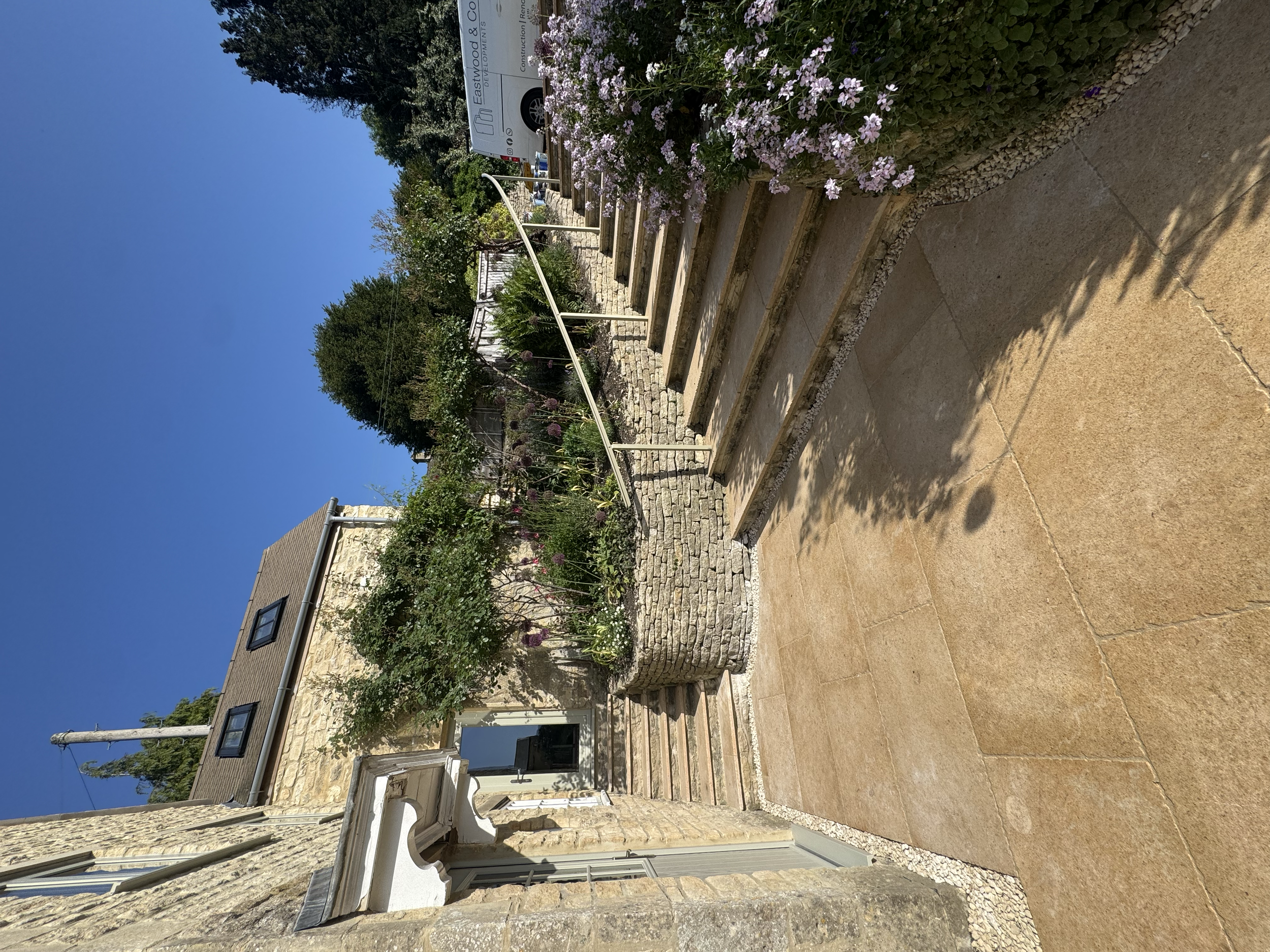 A stone and brick patio area next to a house, with stairs leading up to a garden area filled with bushes and flowering plants. There is a white handrail along the stairs, and a sign on a vehicle in the background reads 'Eastwood & Co.' The house has 