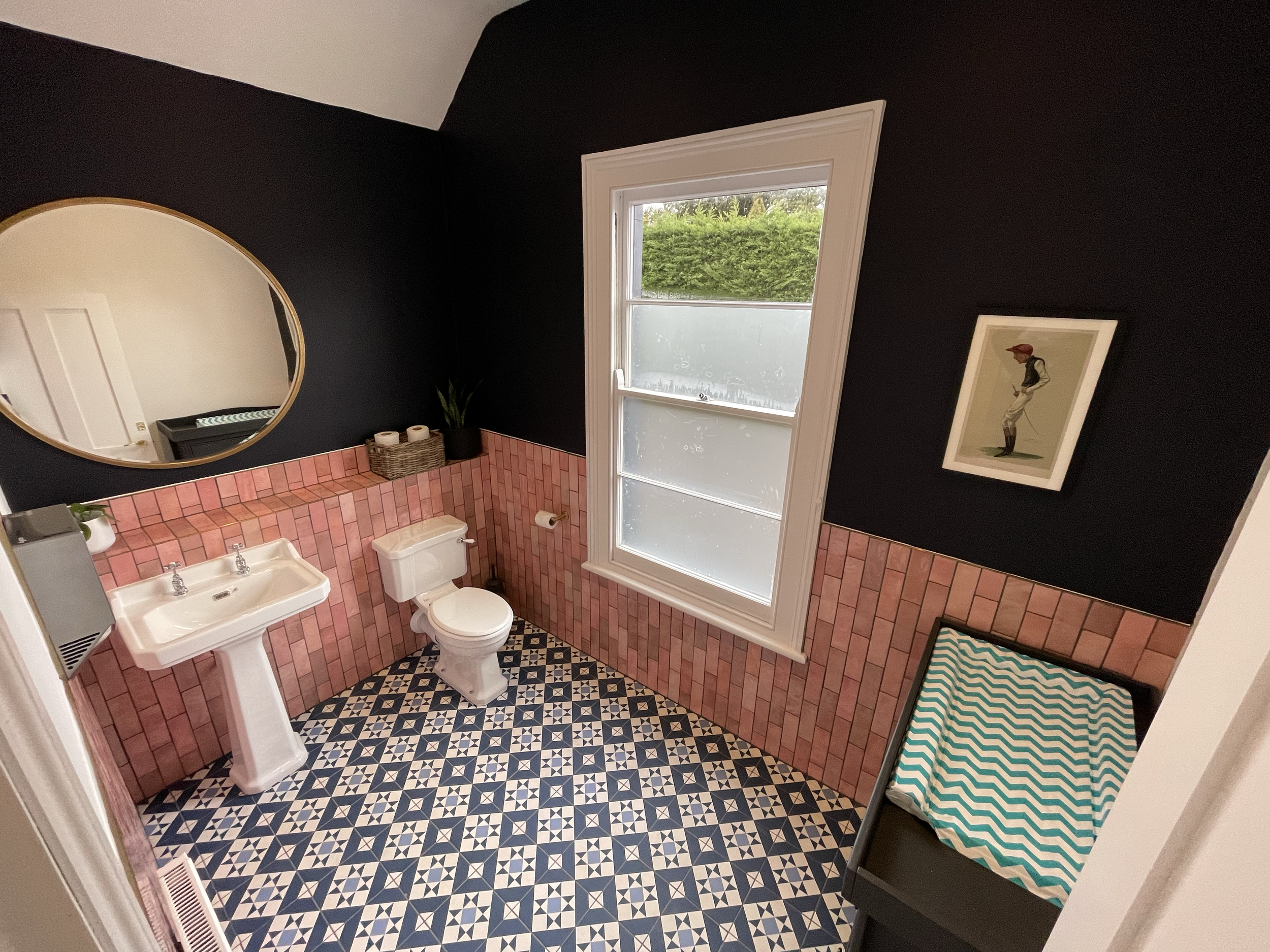 A bathroom with black walls, a window with frosted glass, a white pedestal sink, a toilet, a mirror, a framed picture of a person in vintage clothing, and pink tiled walls. The floor has a black and white geometric pattern, and there is a bench with 