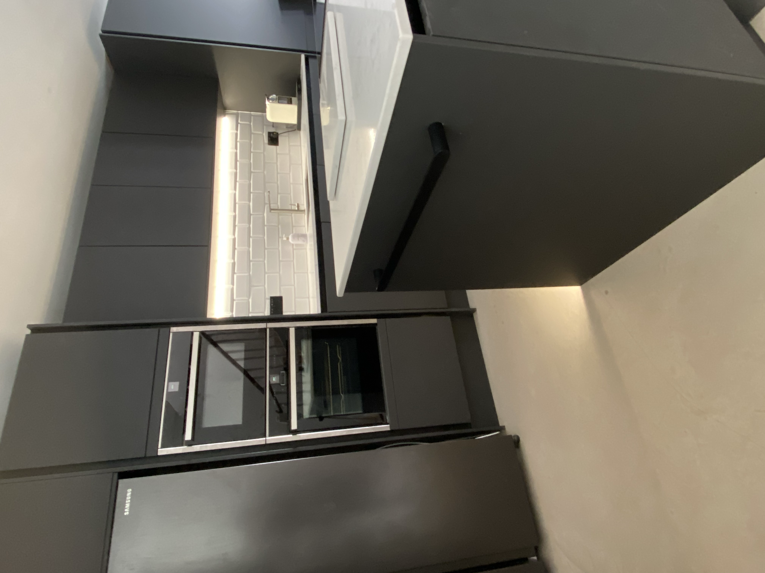 A black kitchen cabinet with a built-in oven and a microwave, dark countertops, and a white backsplash with a geometric pattern.