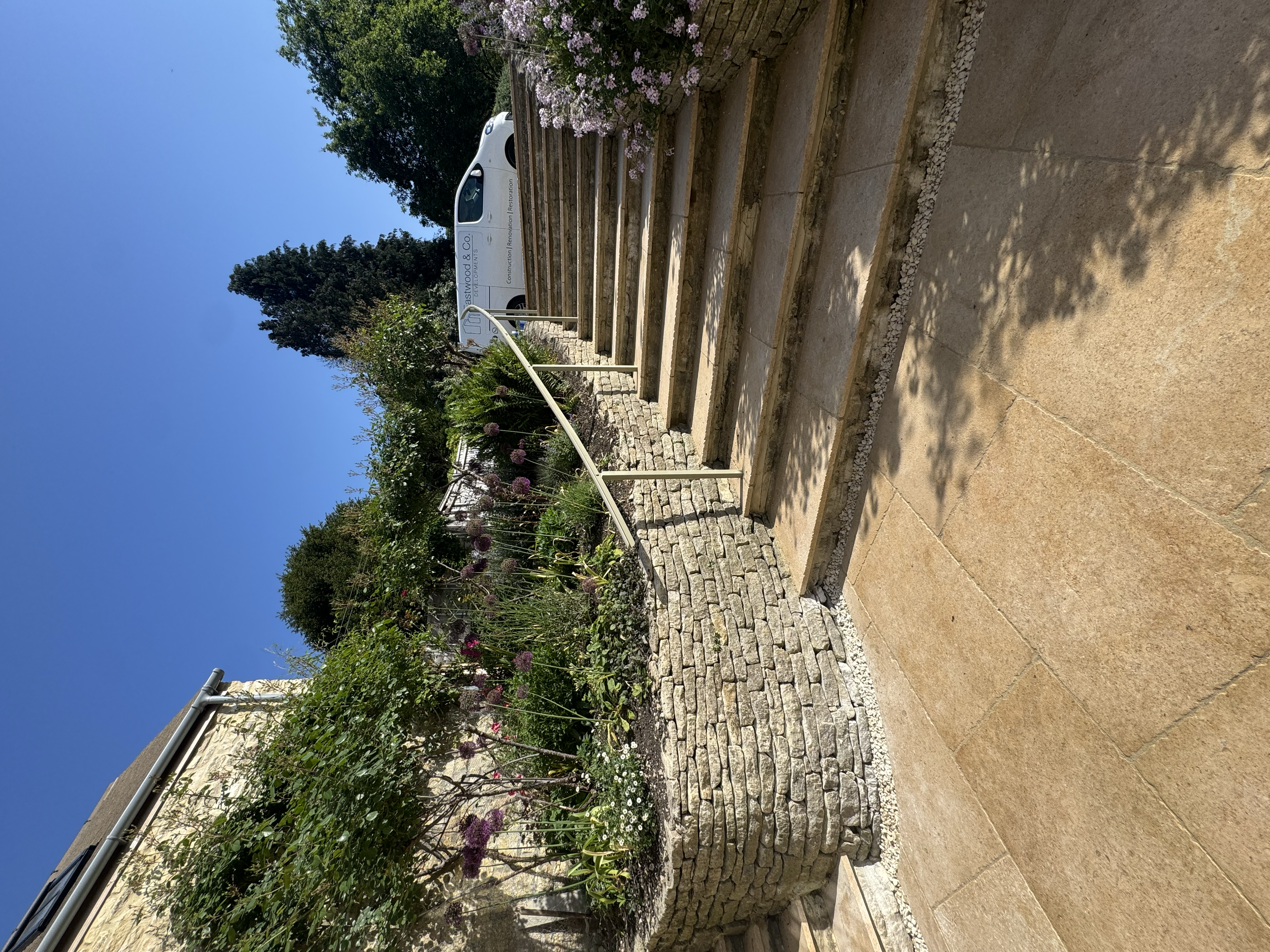 Stone steps leading up to a landscaped garden with plants and flowers, including pink and purple flowers, under a clear blue sky.