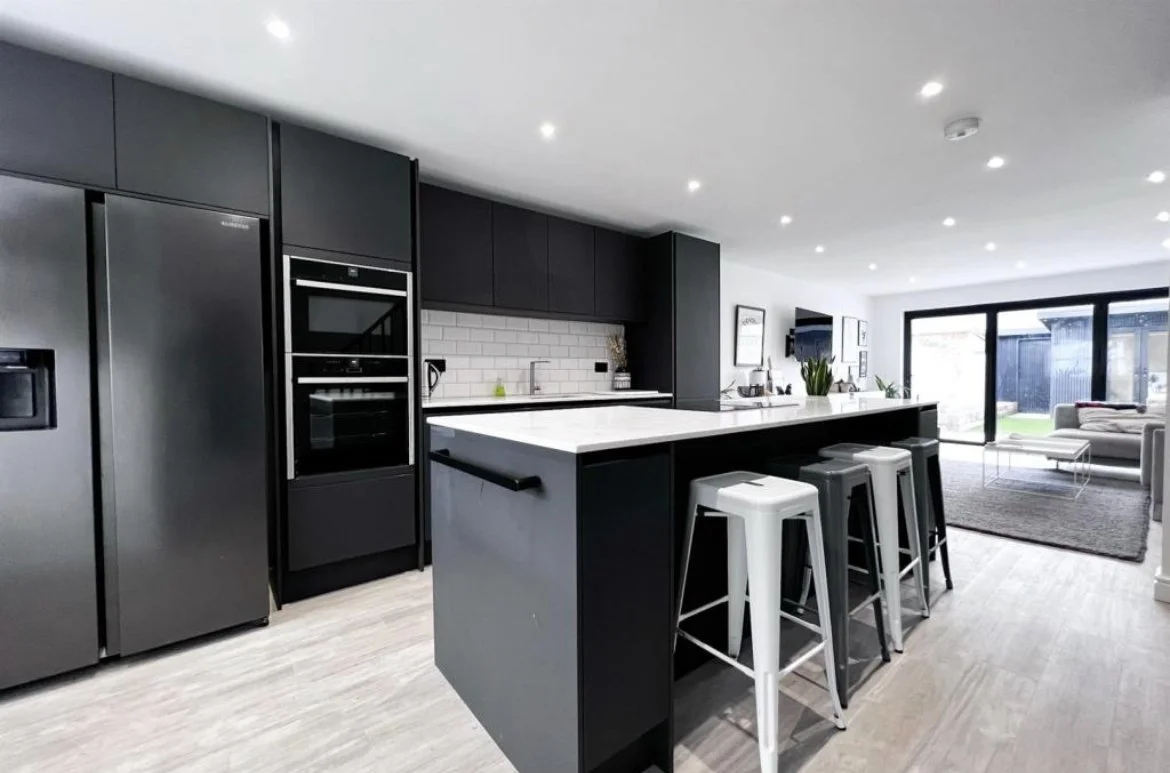 Modern open-concept kitchen with black cabinets, stainless steel appliances, a white island with bar stools, and a living room with a large sliding glass door.