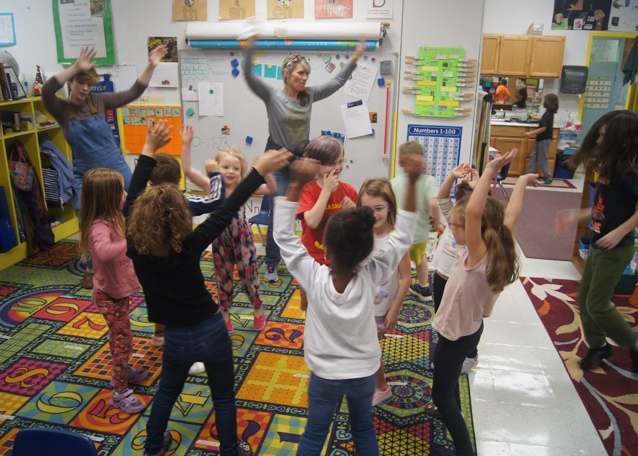 Children in a classroom dancing and playing while a teacher leads them. There are colorful educational posters on the walls and a vibrant patterned carpet.