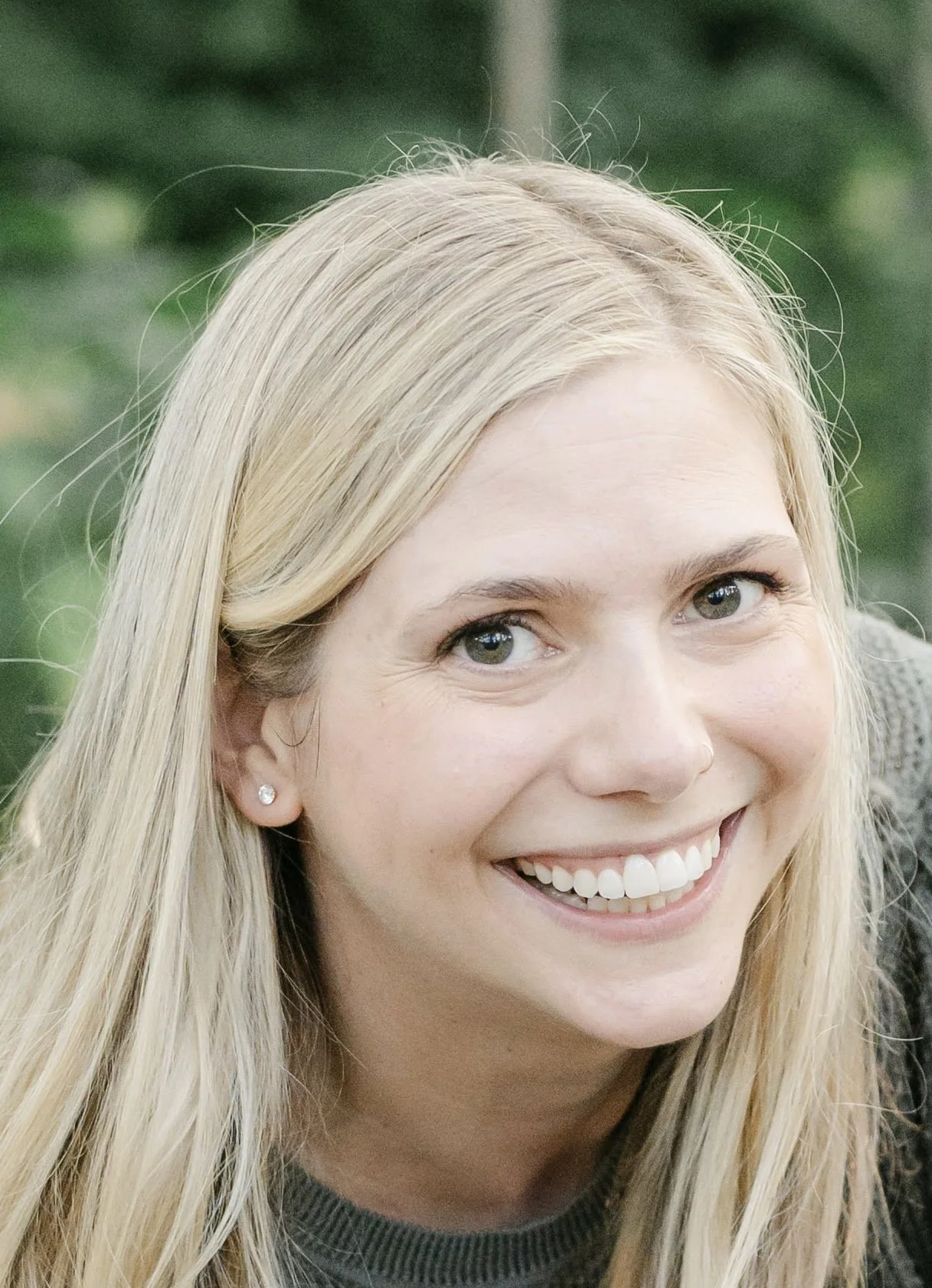 Close-up of a smiling young woman with blonde hair, light skin, wearing a dark gray sweater and small stud earrings, outdoors with blurred green foliage in the background.