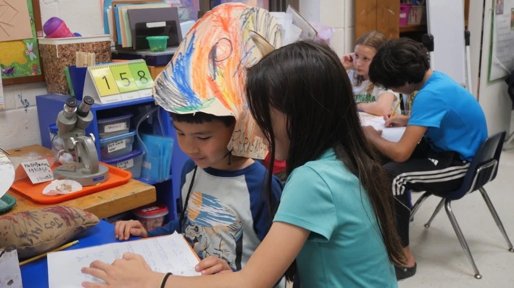 Two children at a table reading a book, with other kids working in the background in a classroom.