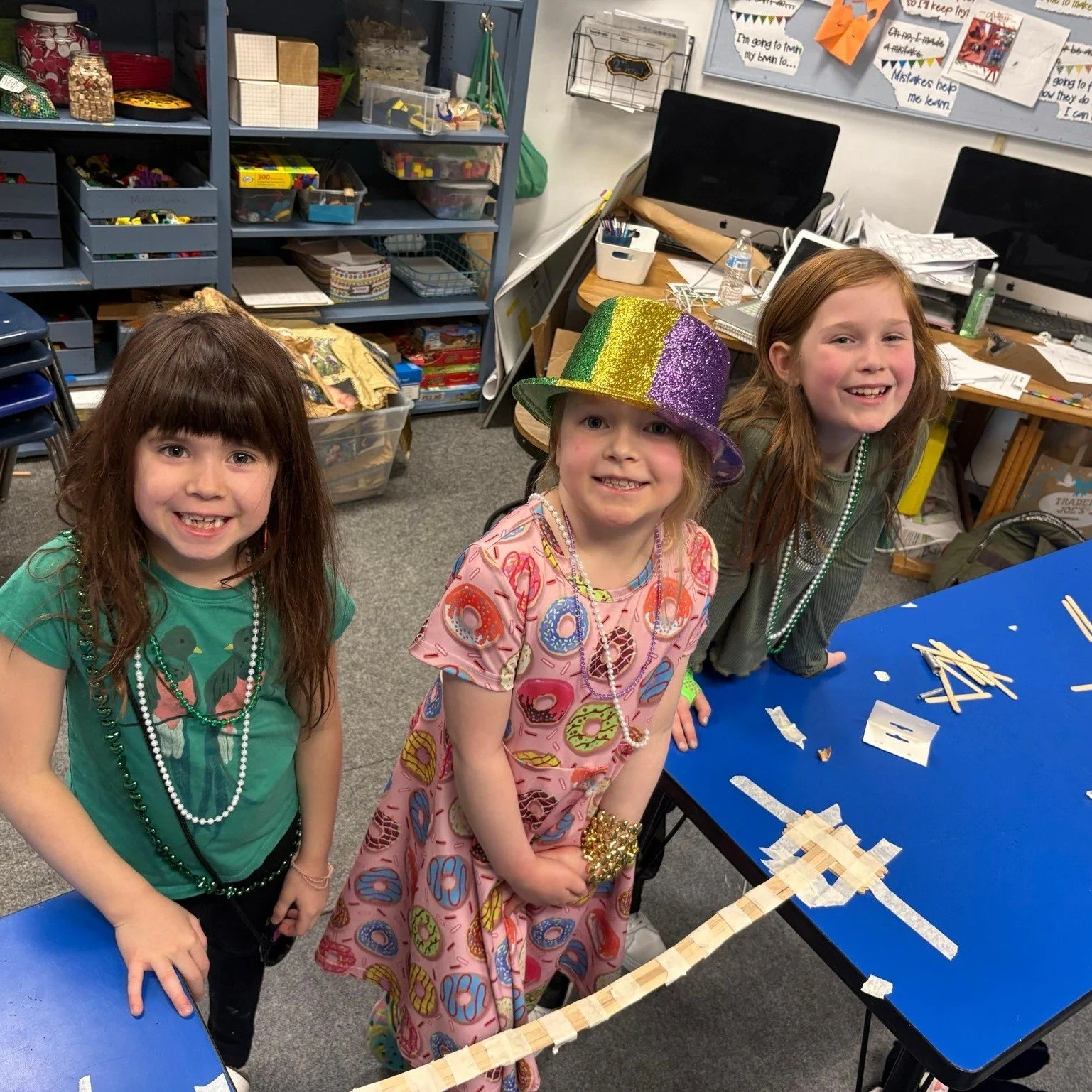Three young girls smiling in a classroom. One girl is wearing a glittery rainbow-colored hat, another is in a green shirt with a dinosaur print, and the third girl is wearing a dress with a donut pattern. They are next to a blue table with wooden sticks and paper pieces, possibly for a craft activity.