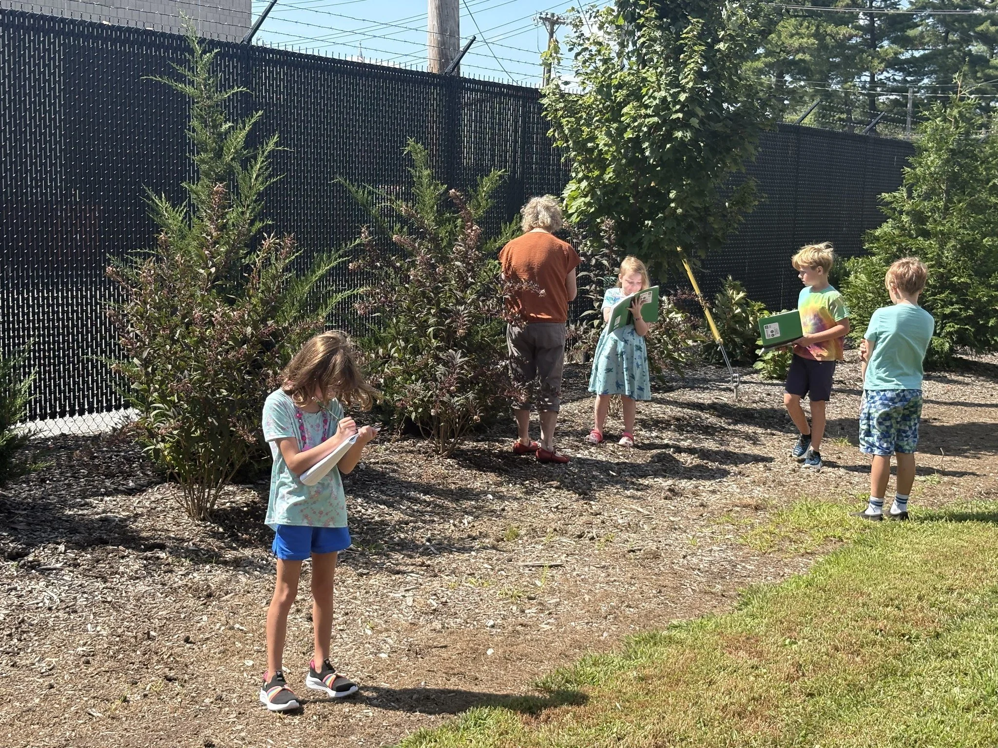 Children and a woman in a garden with bushes, trees, and a tall black fence. Some children are holding notebooks or tablets, and one girl is taking notes, while others are standing or sitting, participating in outdoor activities.