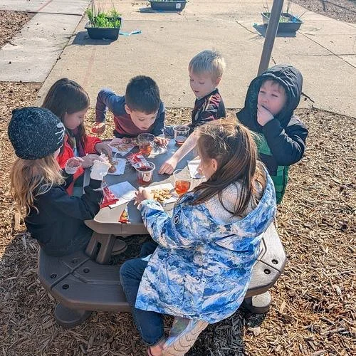 Six children sitting around a table outdoors, eating snacks and drinking from cups.