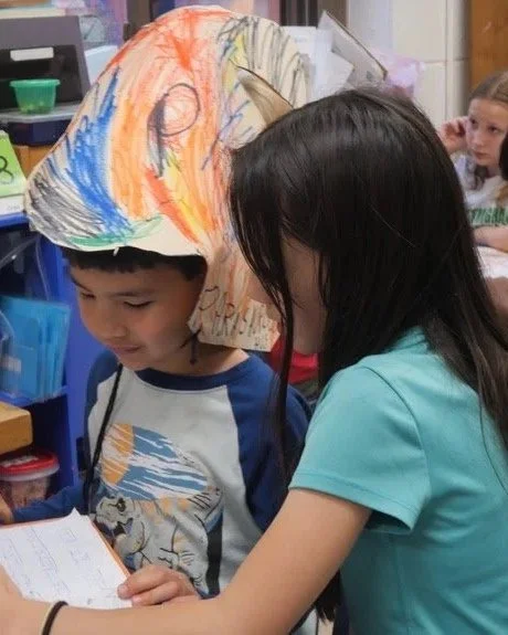 A boy wearing a handmade fish-shaped paper hat, with a girl reading a book together in a classroom setting, with other children visible in the background.