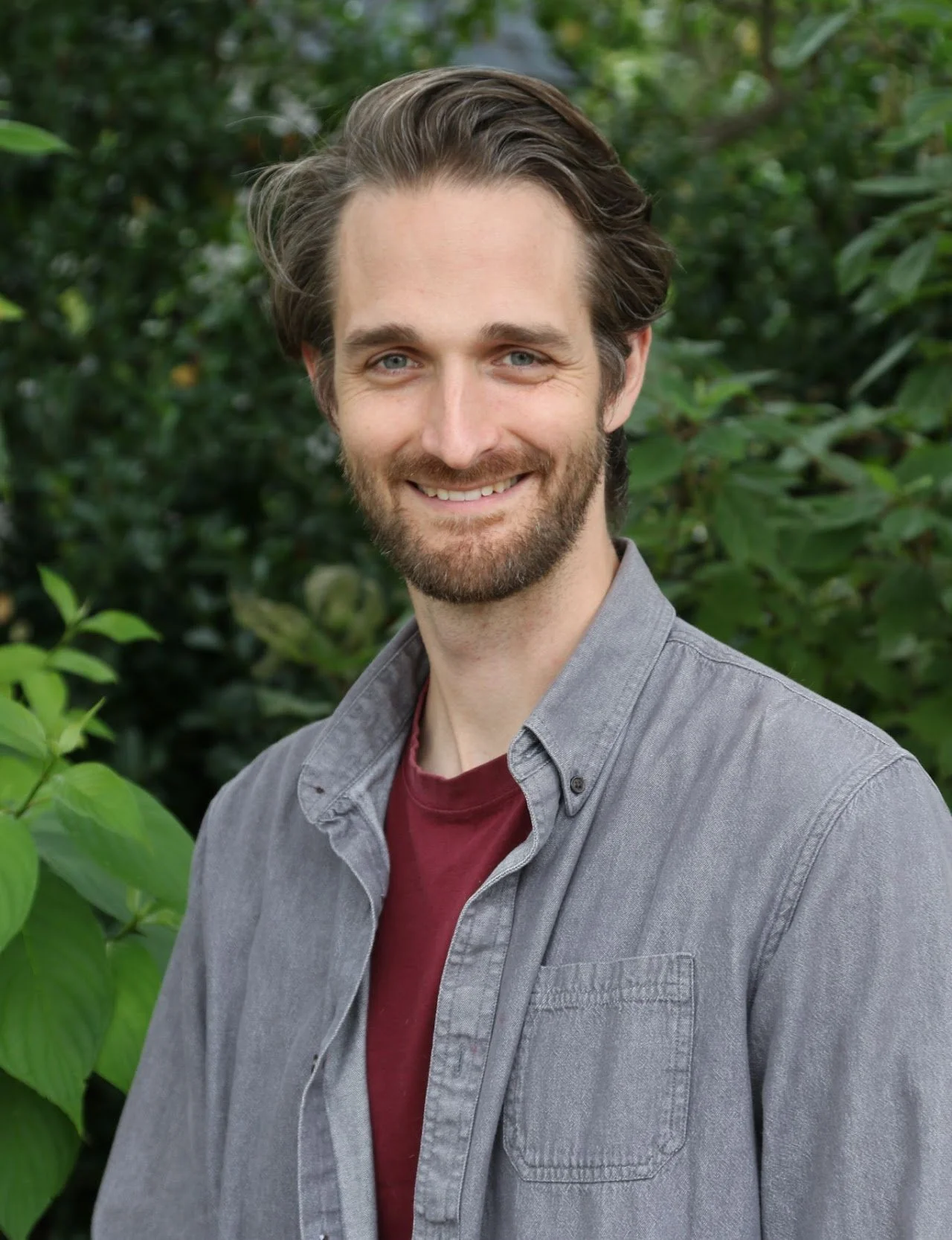 A smiling man with light skin, blue eyes, and brown hair with a beard, standing outdoors in front of green foliage, wearing a gray button-up shirt over a maroon t-shirt.