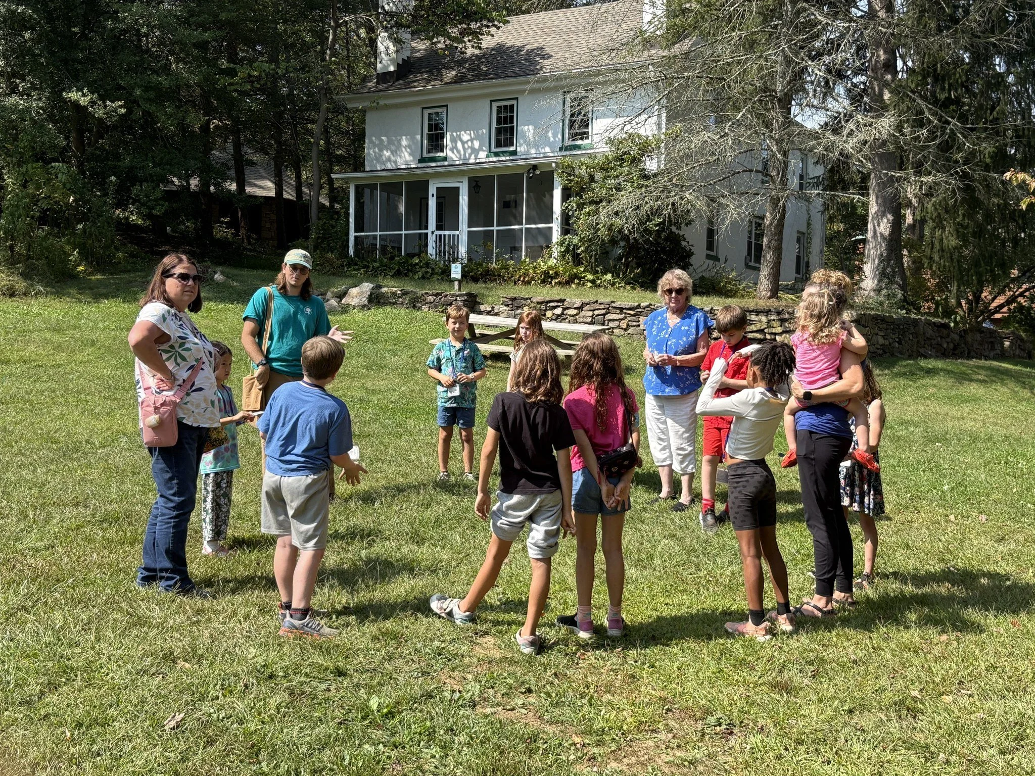 A group of people, including children and adults, gathered on a grassy lawn in front of a white house with a porch, surrounded by trees.