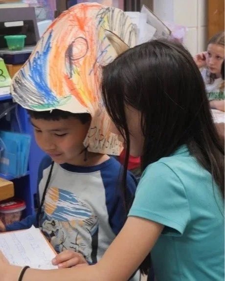 Two children, a boy and a girl, are looking at a book. The boy is wearing a paper helmet decorated with colorful scribbles. The girl has long dark hair and is wearing a light blue shirt. The setting appears to be a classroom.