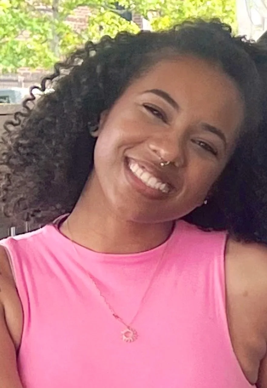 A woman with curly black hair, smiling, wearing a pink sleeveless top and a necklace, outdoors with trees in the background.