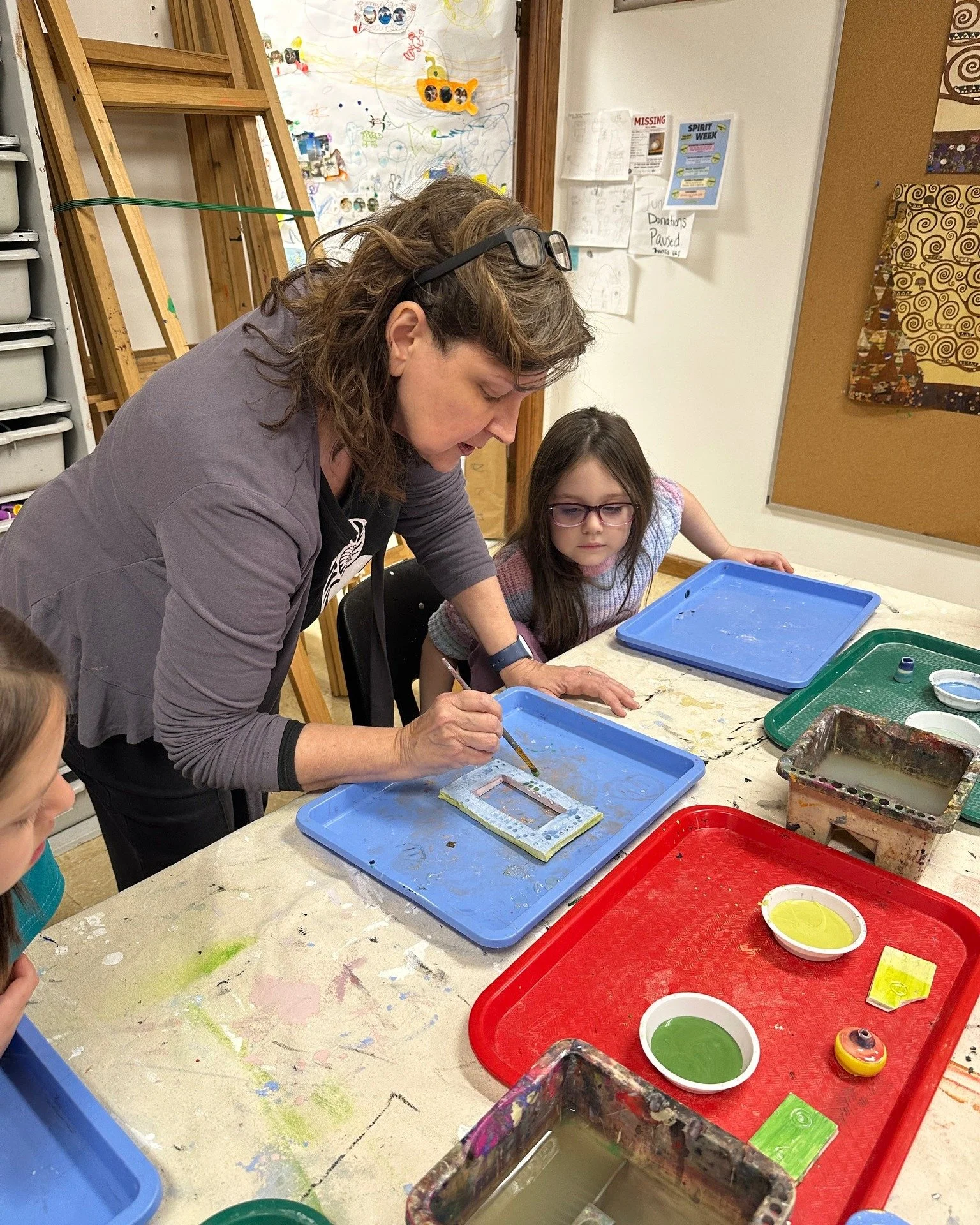 A woman and a young girl engaged in an art project at a table with trays of colorful paints and brushes. The woman is painting a design on a piece of material while the girl observes. The table and background are cluttered with art supplies and colorful artwork.