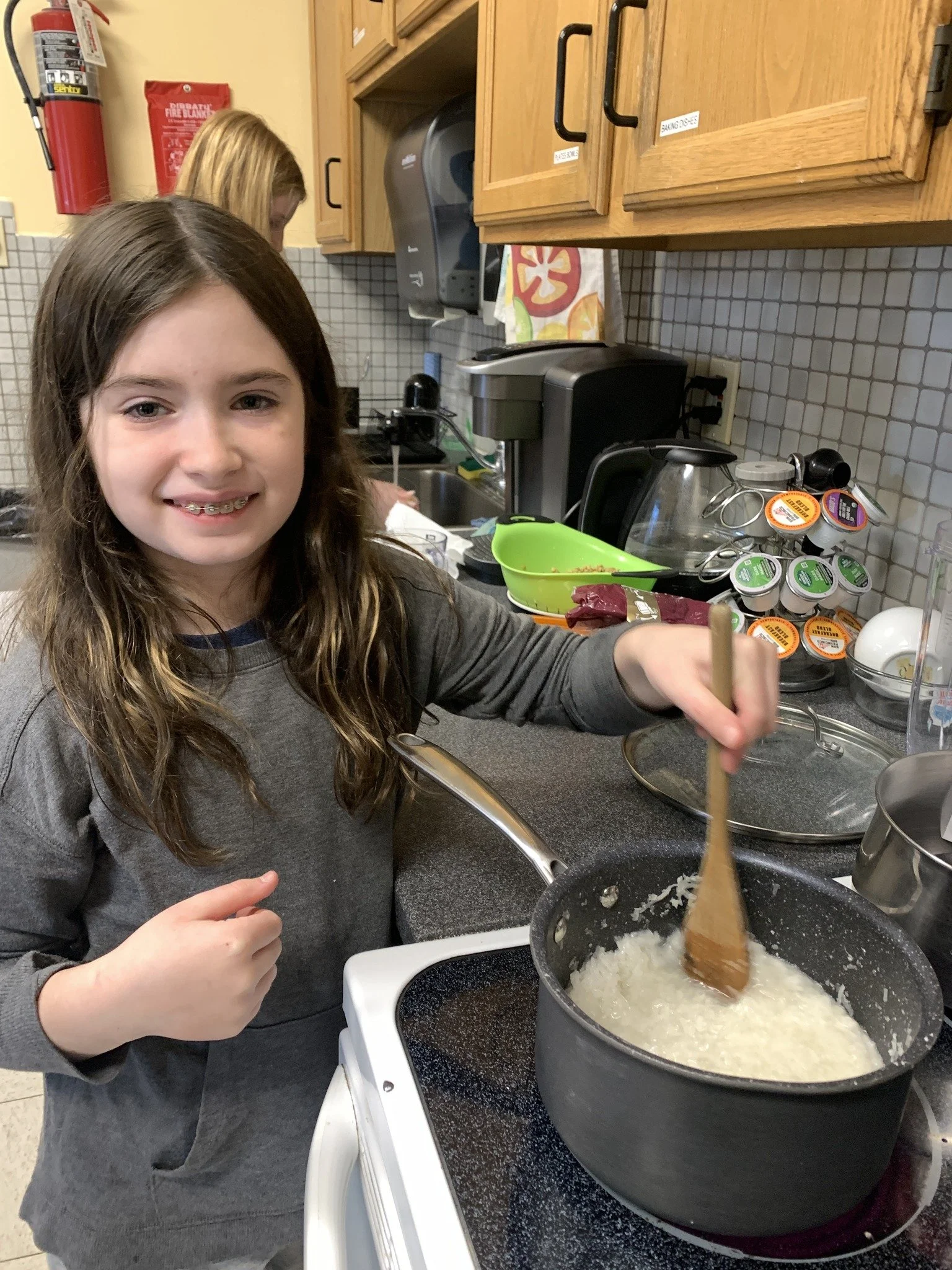 A young girl with brown hair and braces stirring rice in a pot on a stove, with a smiling girl in the background in a kitchen.