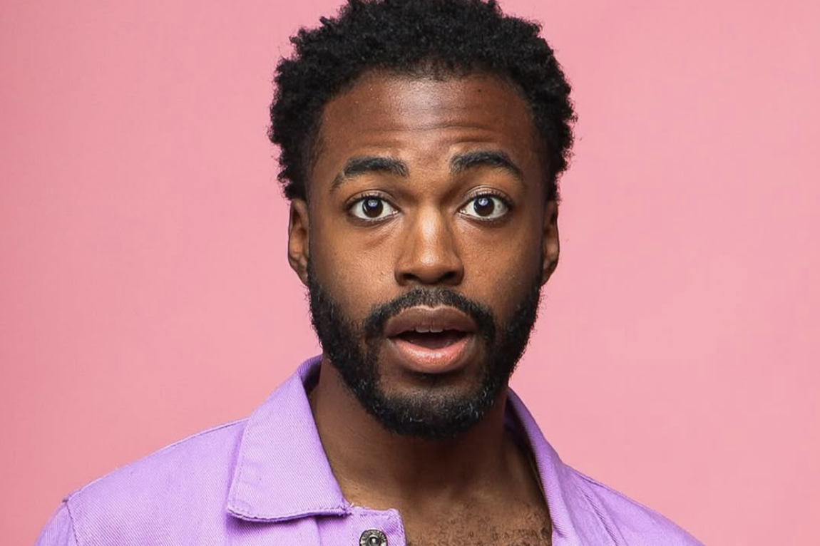 A man with dark curly hair and a beard, wearing a purple shirt, looking surprised against a pink background.