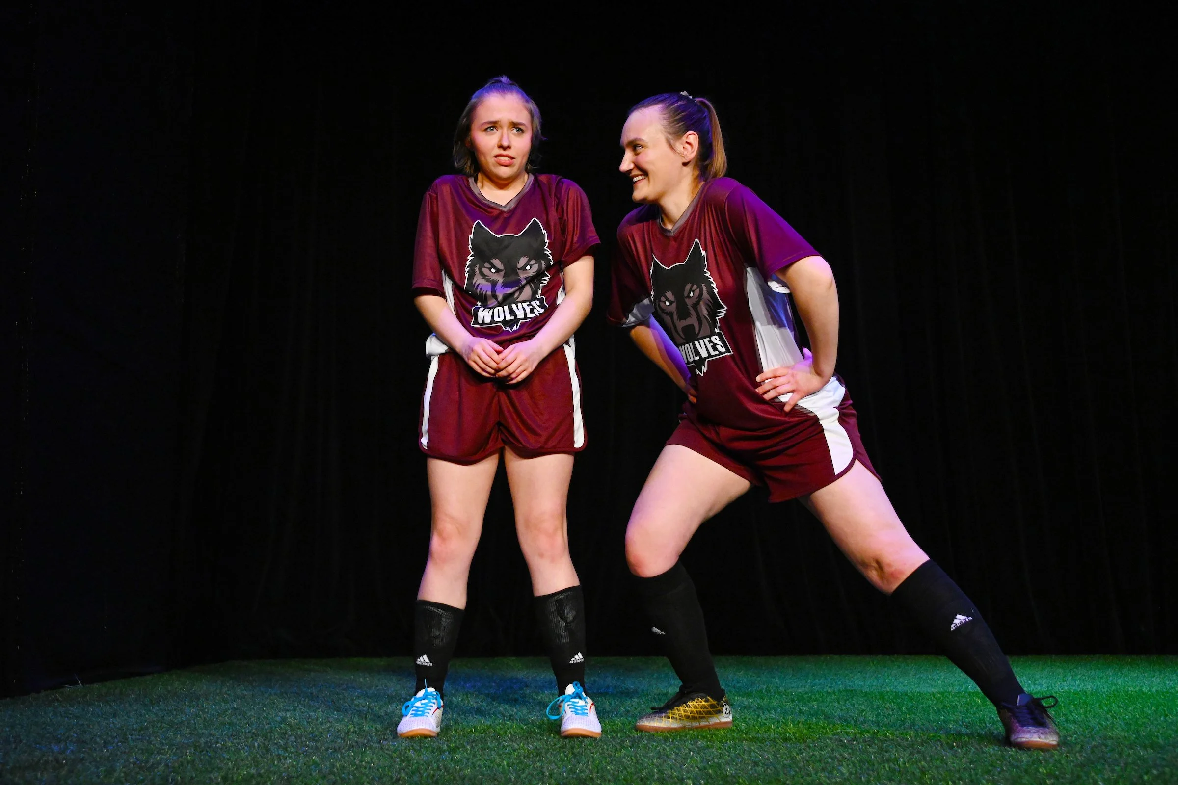 Two young women wearing matching maroon sports uniforms with wolf and 'Wolves' logos, one with a surprised expression and the other smiling, engaging in a theatrical performance on stage with a black background.