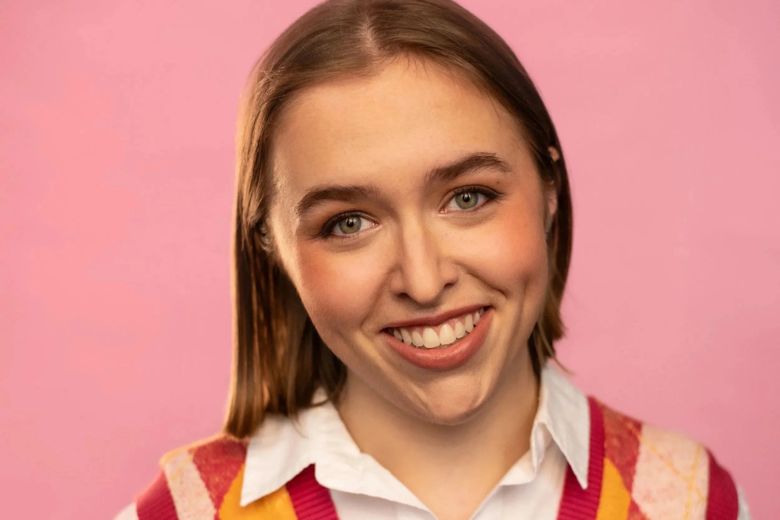 Close-up of a smiling young woman with light brown hair and blue eyes, wearing a white collared shirt and a red and yellow argyle sweater vest, against a pink background.