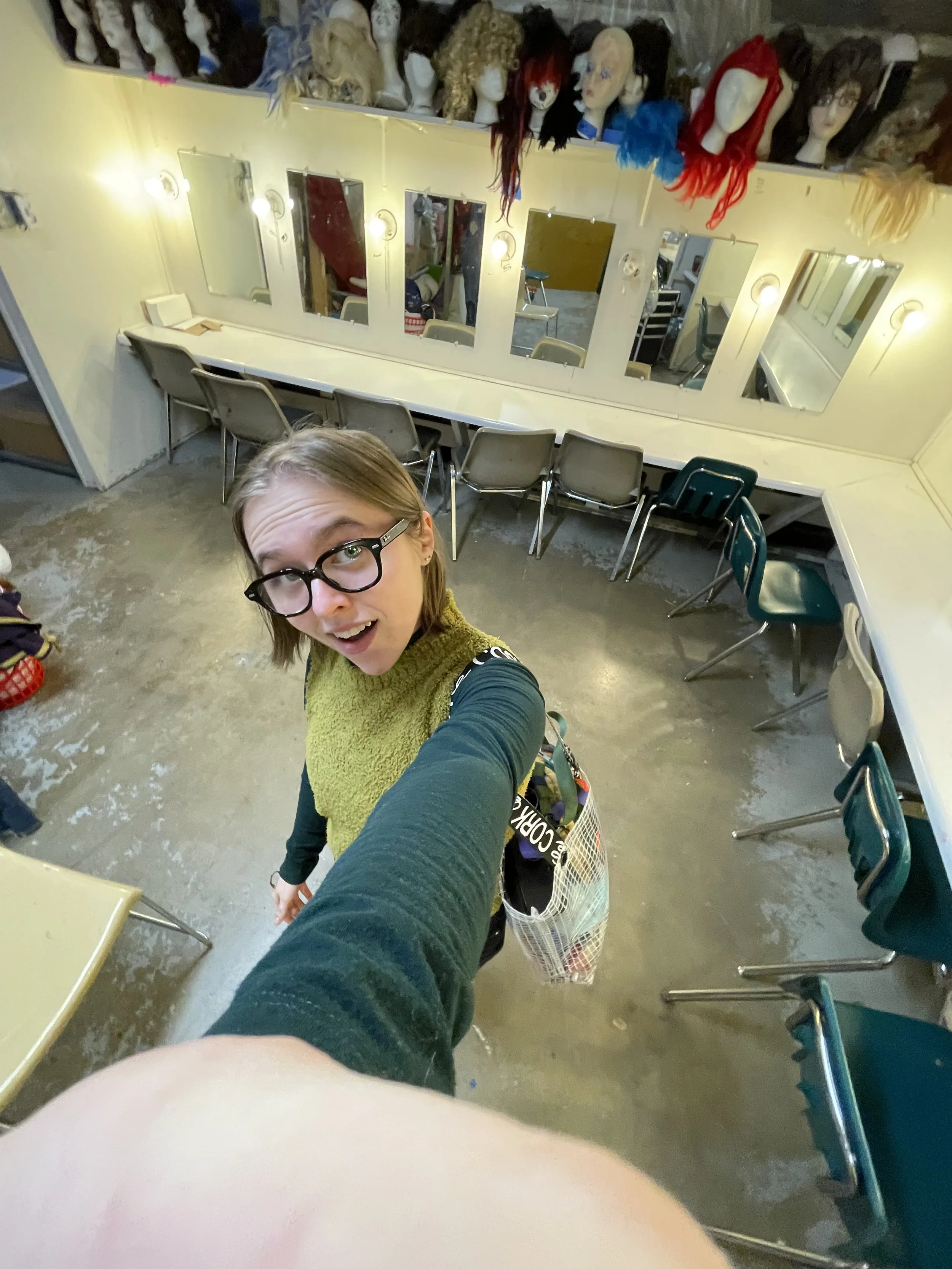 A young woman with glasses and a yellow-green sweater is taking a selfie in a dressing room with multiple mirrors, wigs on display on the wall above, and various chairs around a white counter.