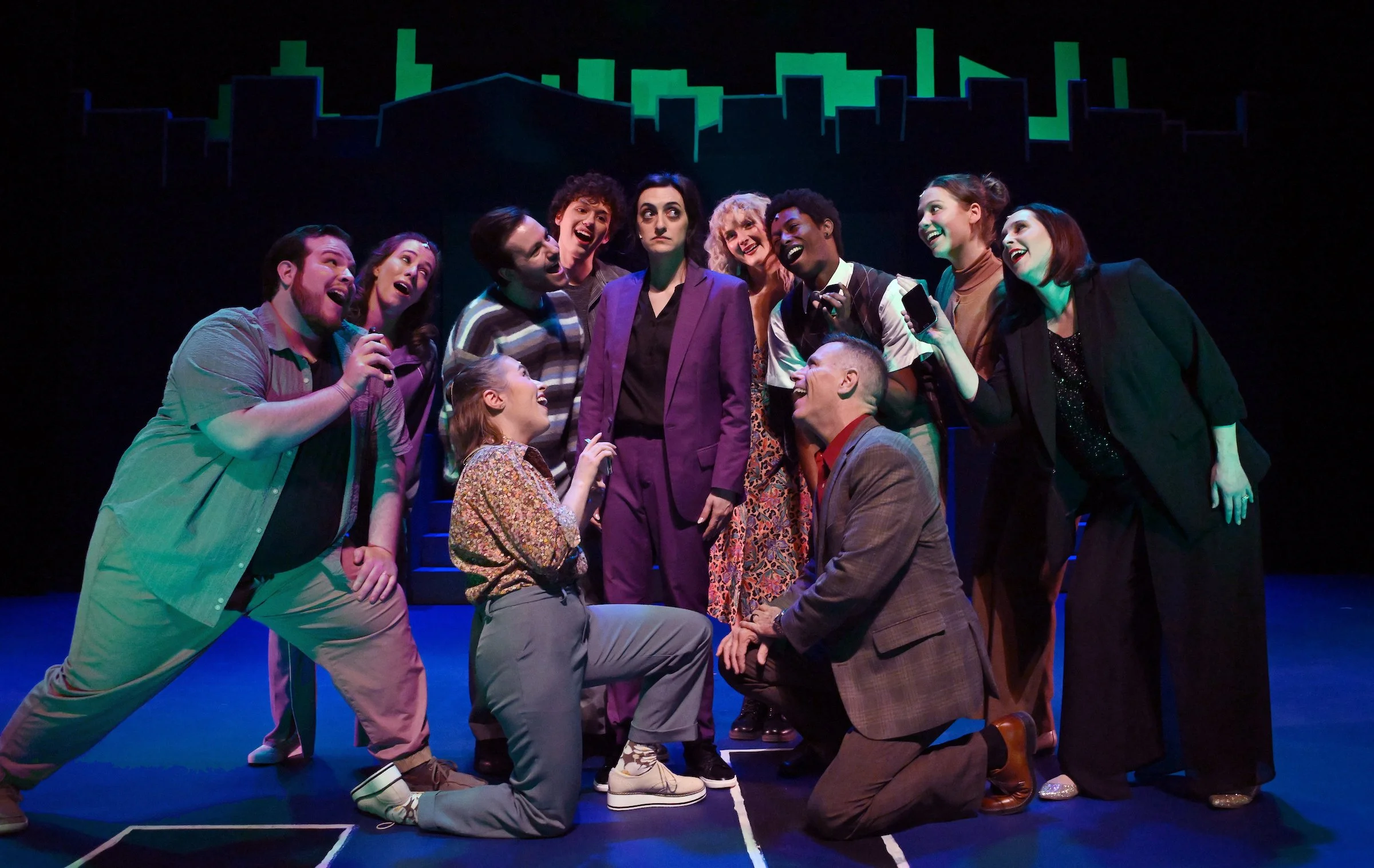 A theatrical cast on stage, with some actors kneeling and others standing, engaged in a scene with expressive facial expressions and diverse costumes. The background features a stylized city skyline silhouette in green.