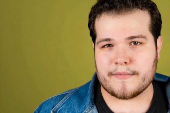 Head and shoulders portrait of a young man with short dark hair, light skin, and facial hair, wearing a black shirt and a denim jacket, against a green background.