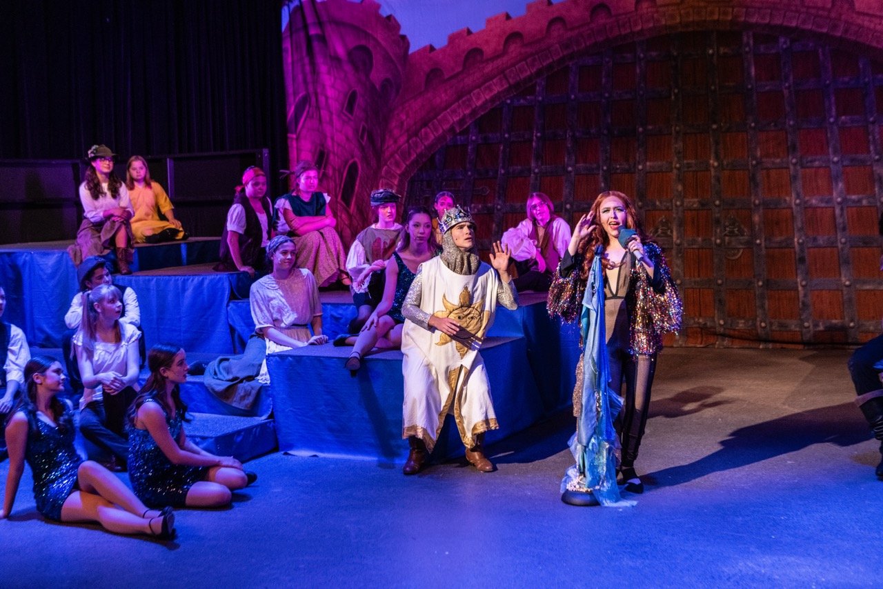 Children performing on stage in a theatrical play, some sitting and some standing, with a woman singing into a microphone and dressed in a colorful, sparkly costume, with a castle backdrop.