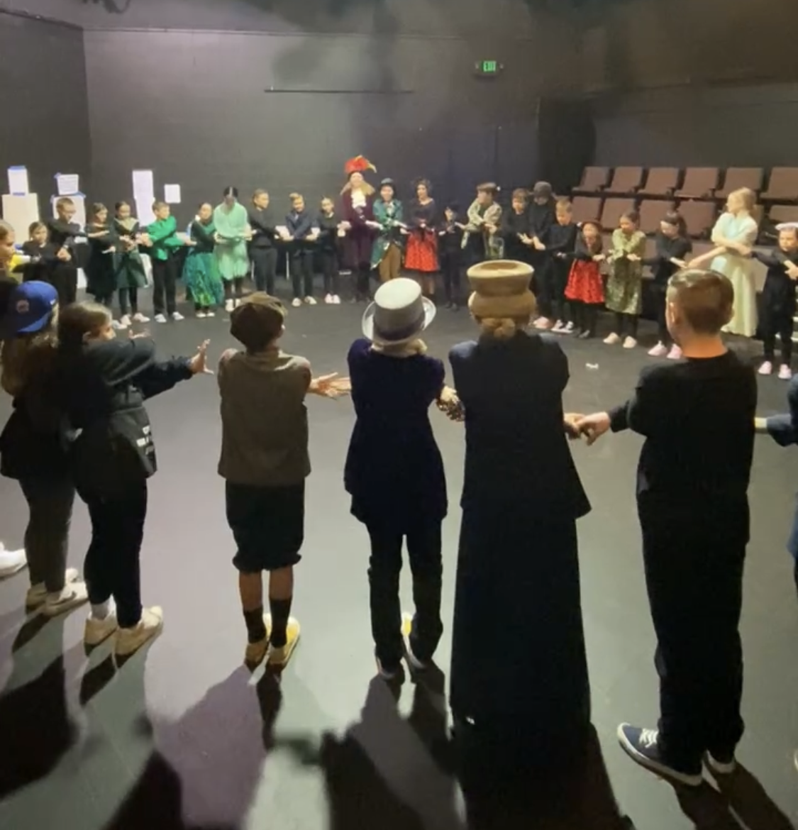 Children in a circle holding hands, dressed in costumes for a show, in a dimly lit indoor space.